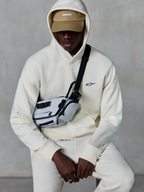 Sudadera con capucha Alpinestars Essentials Ageless, estilo streetwear casual, color blanco roto, pequeño logo negro bordado en el pecho, capucha puesta sobre una gorra color tostado con marca ASTARS, combinada con una bandolera gris claro, ropa lifestyle premium