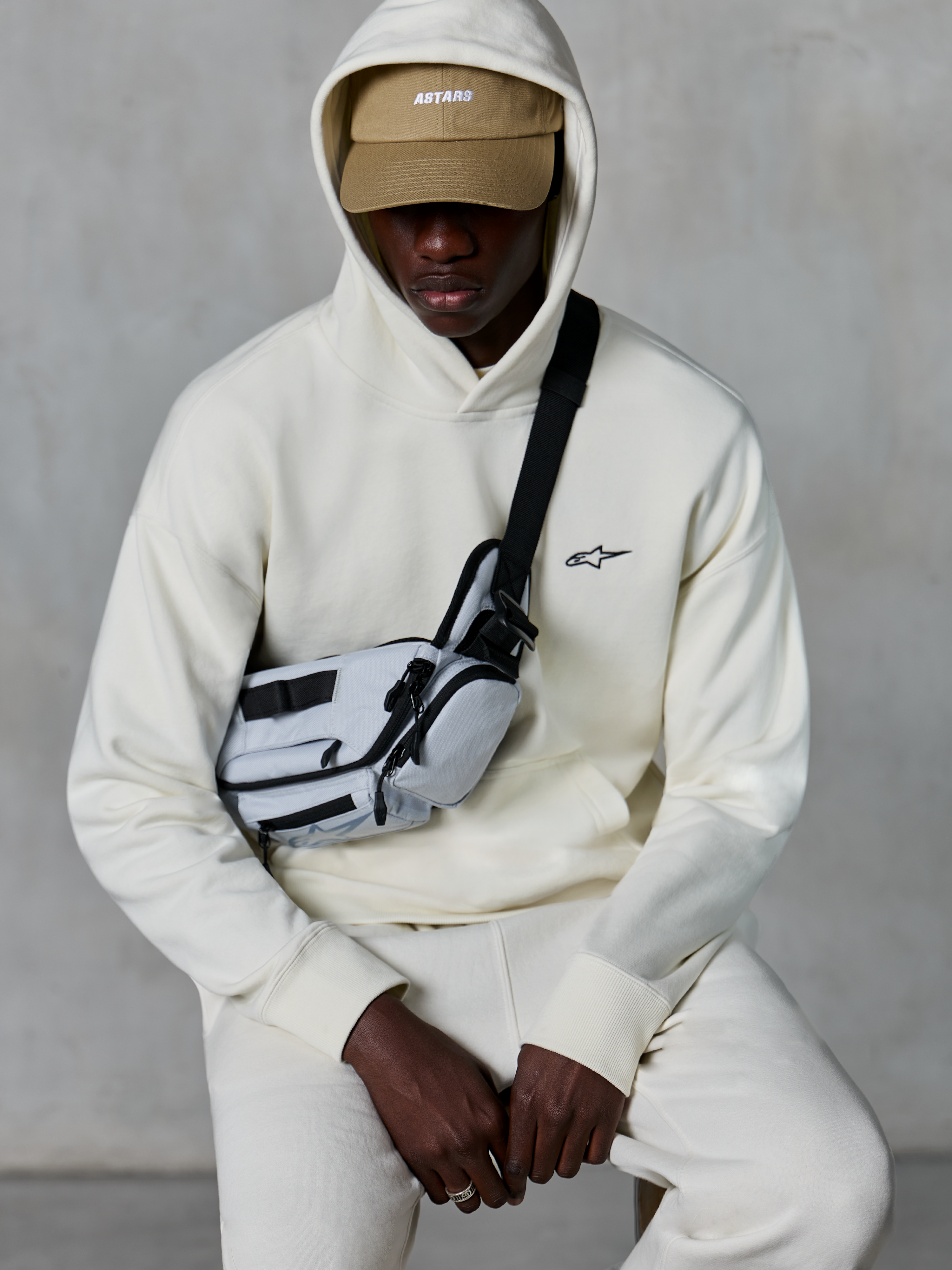 Sudadera con capucha Alpinestars Essentials Ageless, estilo streetwear casual, color blanco roto, pequeño logo negro bordado en el pecho, capucha puesta sobre una gorra color tostado con marca ASTARS, combinada con una bandolera gris claro, ropa lifestyle premium