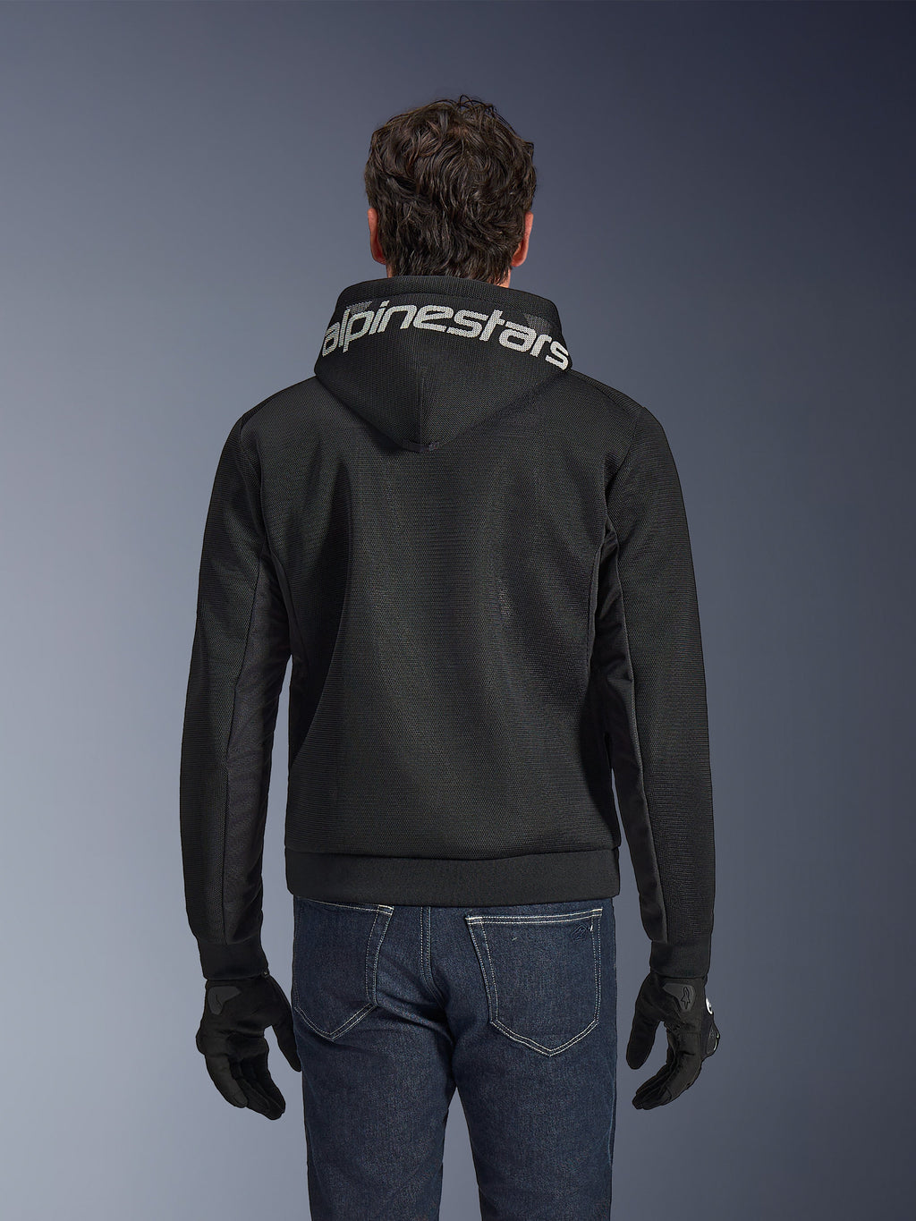 Alpinestars Chrome Superairflow Hoodie, sudadera de motociclismo negra, vista trasera con el logotipo de Alpinestars en blanco en la capucha, construcción de malla técnica 3D, combinada con vaqueros azul oscuro y guantes negros, prenda de conducción urbana ventilada.