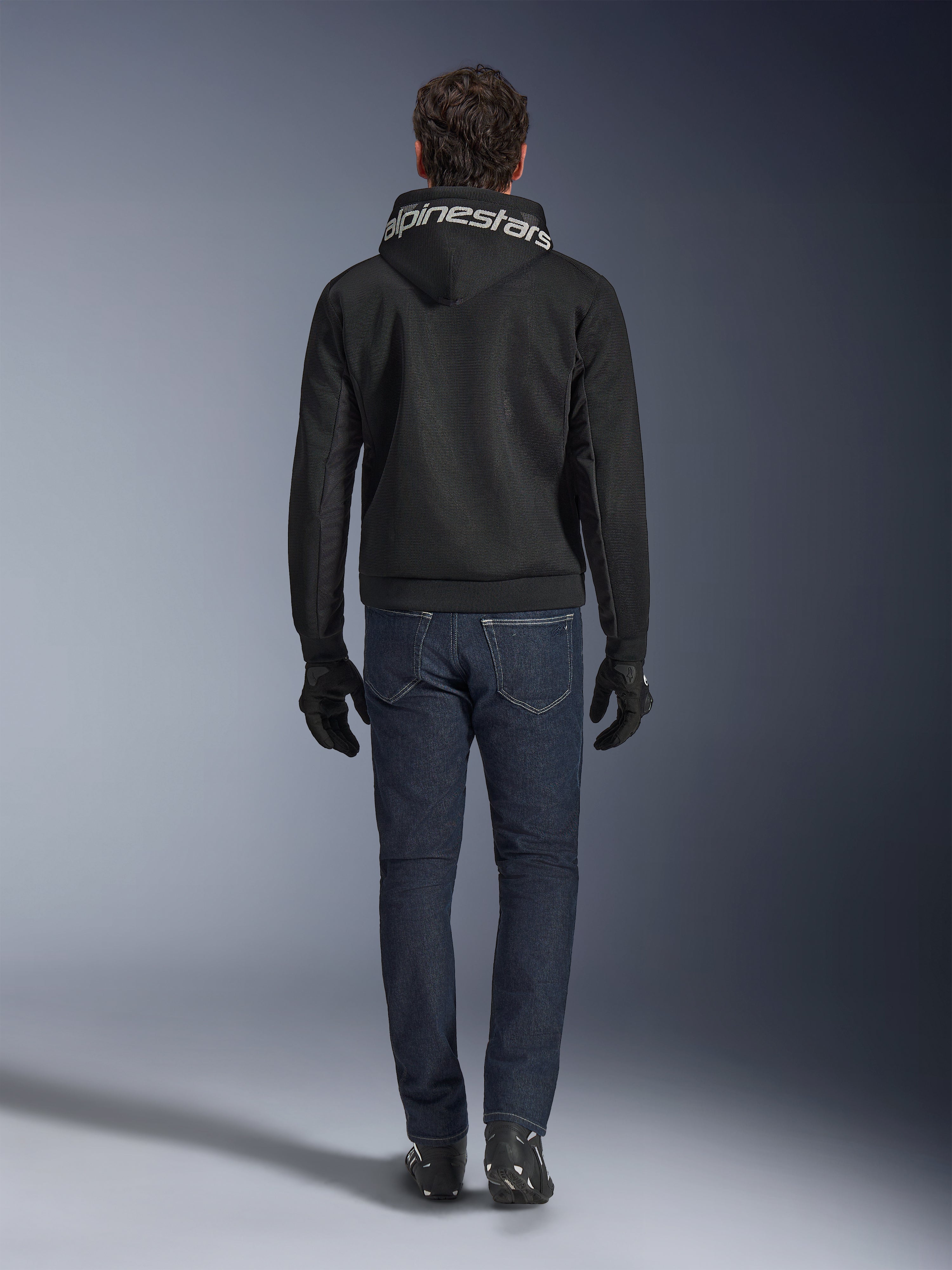 Sudadera con capucha Alpinestars Chrome Superairflow, sudadera de moto negra con logotipo blanco en la capucha, vista desde atrás, combinada con vaqueros azul oscuro, guantes negros y botas blancas y negras, equipamiento de conducción urbana y deportiva