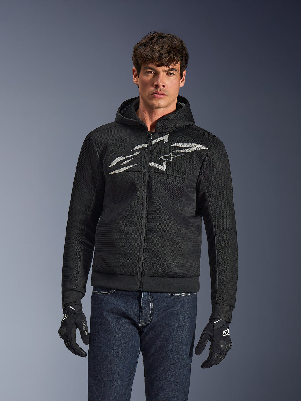 Alpinestars Chrome Superairflow Hoodie, sudadera de moto negra con cremallera y un gran logotipo blanco en el pecho, construcción de malla ventilada, combinada con vaqueros azules y guantes negros, ropa deportiva para conducción urbana