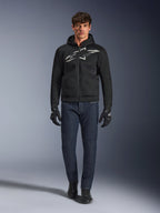 Motociclista con la sudadera con capucha Alpinestars Chrome Superairflow en negro y gris, combinada con vaqueros azul oscuro, guantes de moto negros y calzado negro. La sudadera presenta un gran logotipo estilizado en el pecho y una textura de malla 3D para un alto rendimiento en temperaturas elevadas.