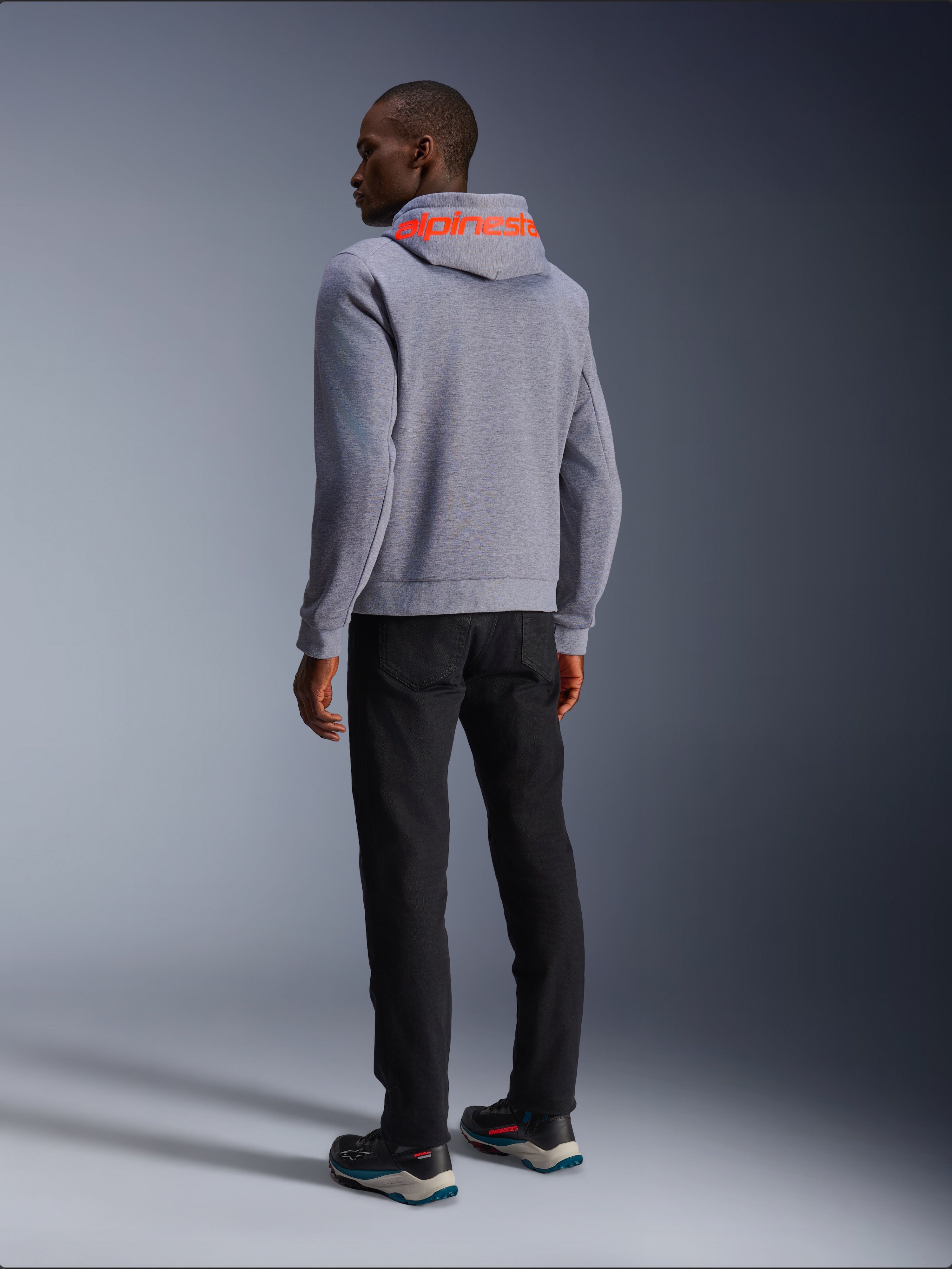 Alpinestars Chrome V2 Sport Hoodie in Gray Heather, back view showing a large Red Fluo Alpinestars wordmark across the hood, worn by a model with black pants and dark riding shoes, casual urban motorcycle apparel