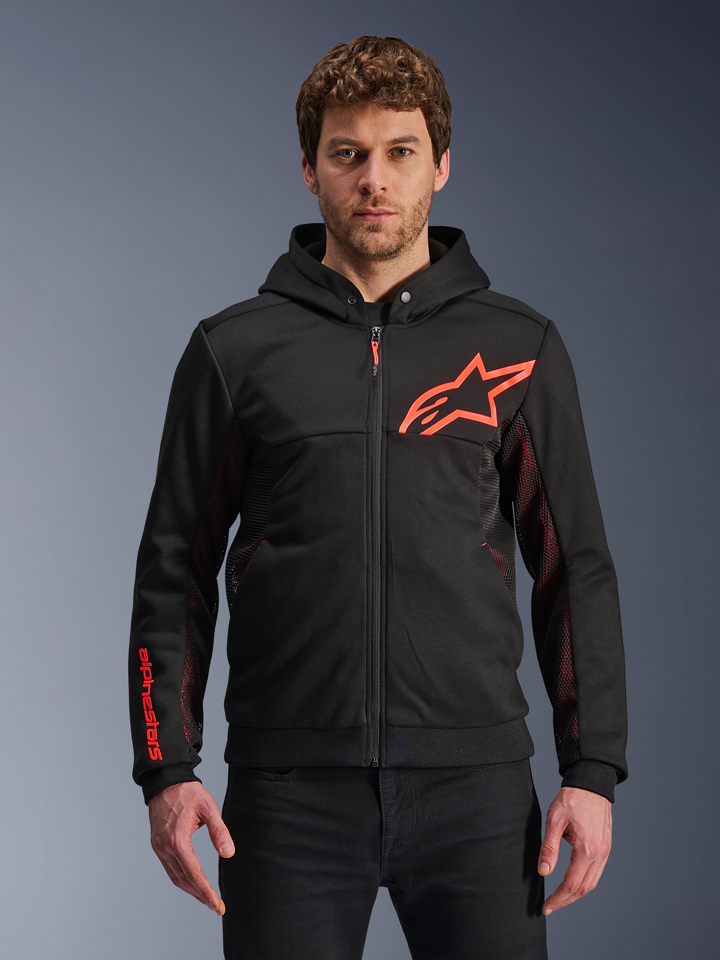 Alpinestars Chrome Air Sport Hoodie, sudadera negra con cremallera y capucha con logotipo de estrella en rojo flúor y detalles en las mangas, paneles laterales de malla roja para ventilación, equipamiento de conducción urbana casual usado por un modelo
