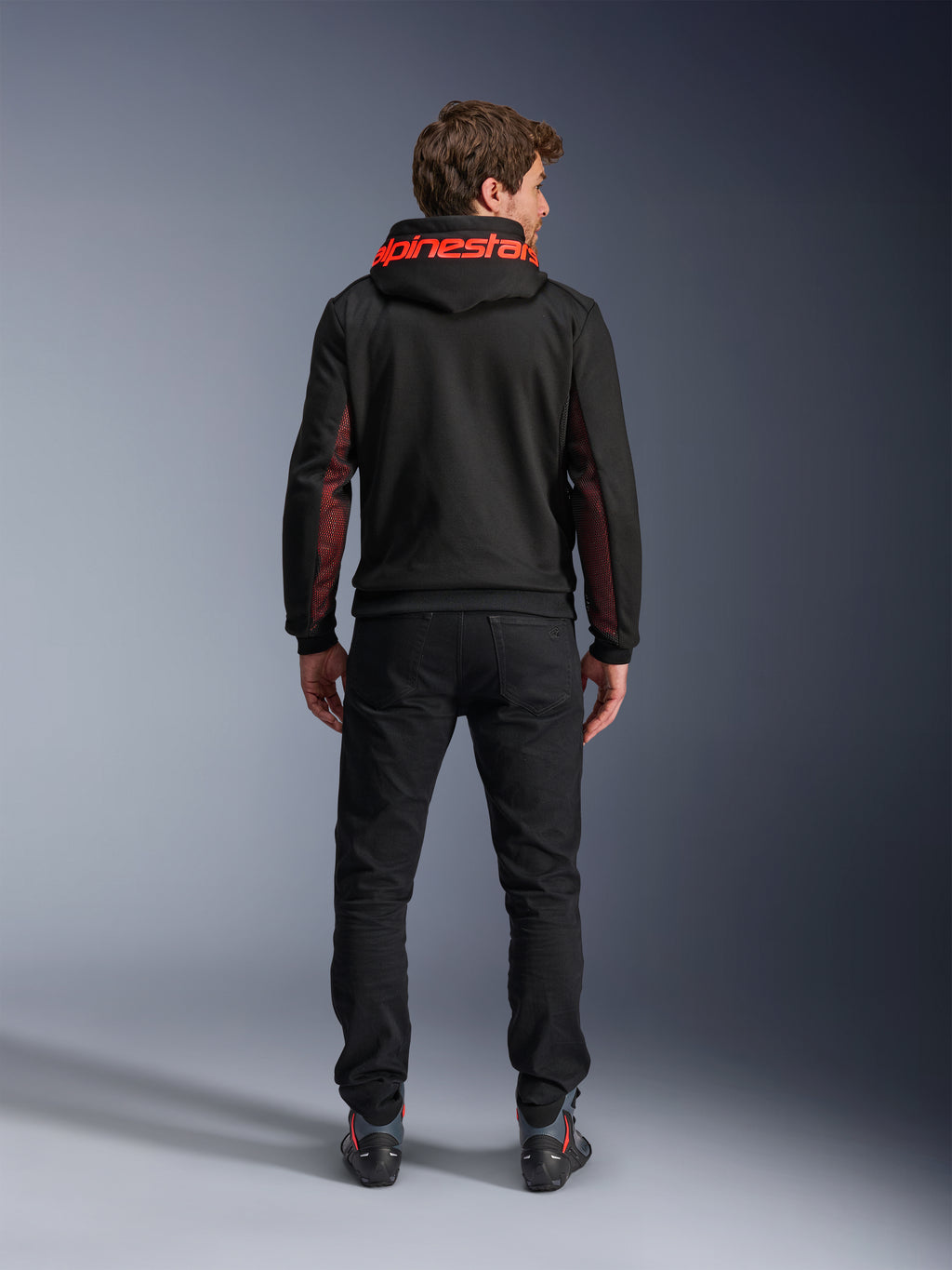 Alpinestars Chrome Air Sport Hoodie, negro y rojo fluo, vista posterior de un hombre con chaqueta de moto casual, con la marca Alpinestars en rojo en la capucha y paneles de malla rojos en las mangas, combinada con pantalones negros.