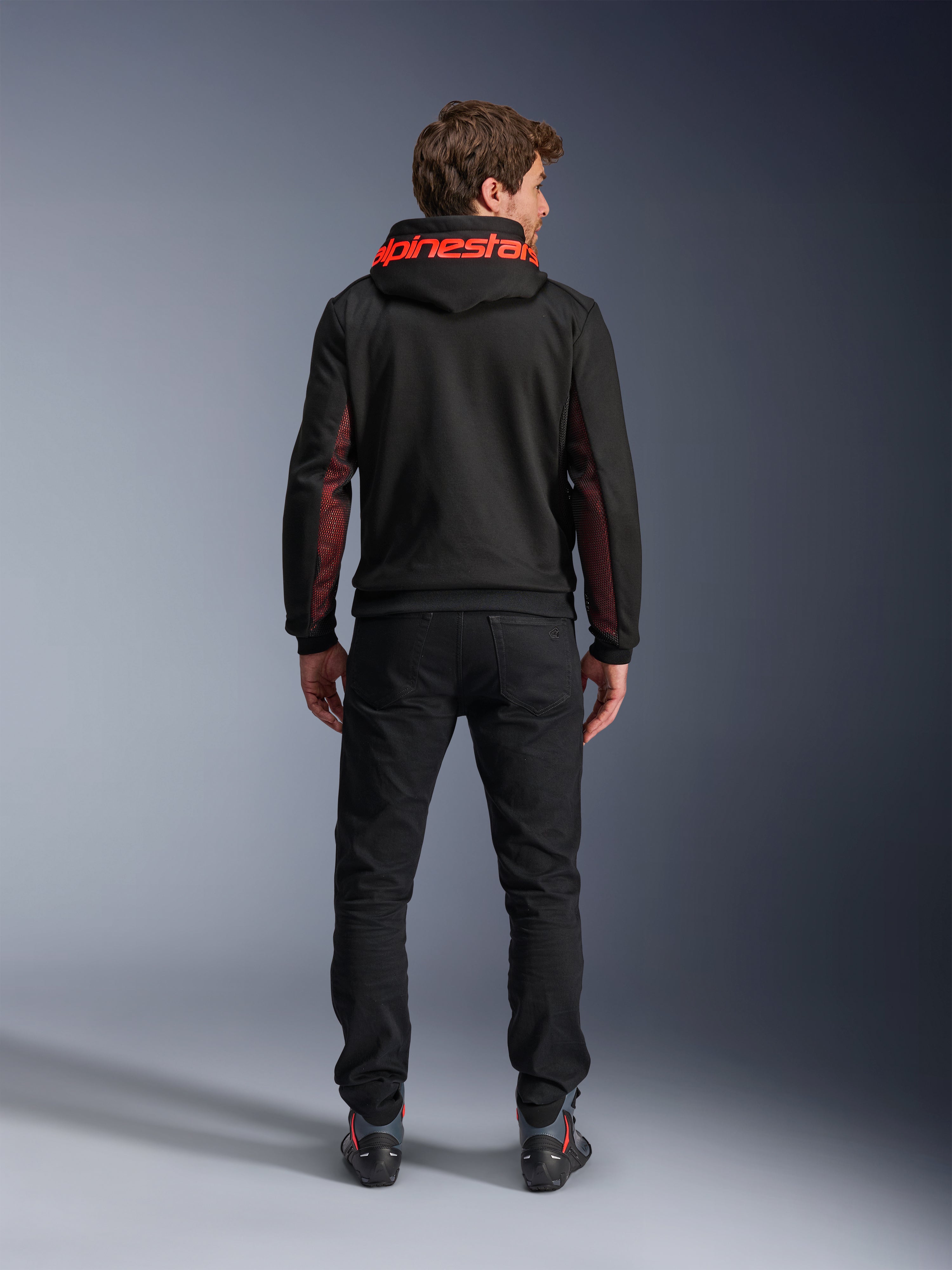 Alpinestars Chrome Air Sport Hoodie, negro y rojo fluo, vista posterior de un hombre con chaqueta de moto casual, con la marca Alpinestars en rojo en la capucha y paneles de malla rojos en las mangas, combinada con pantalones negros.