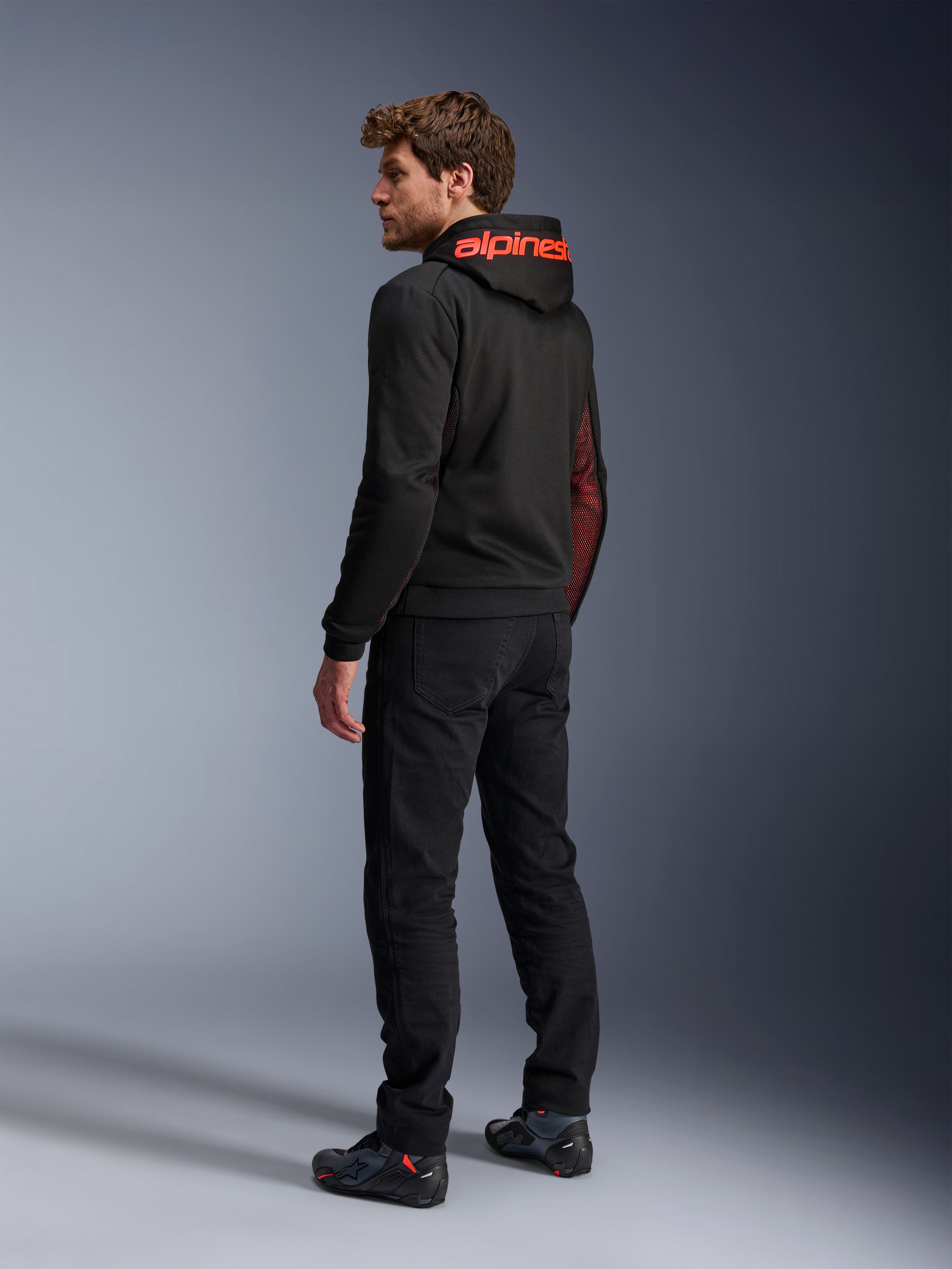 Sudadera Alpinestars Chrome Air Sport, sudadera de moto urbana, negro y rojo flúor, vista trasera que muestra la marca Alpinestars en rojo en la capucha y los paneles laterales de malla, usada con pantalones de moto negros y calzado técnico