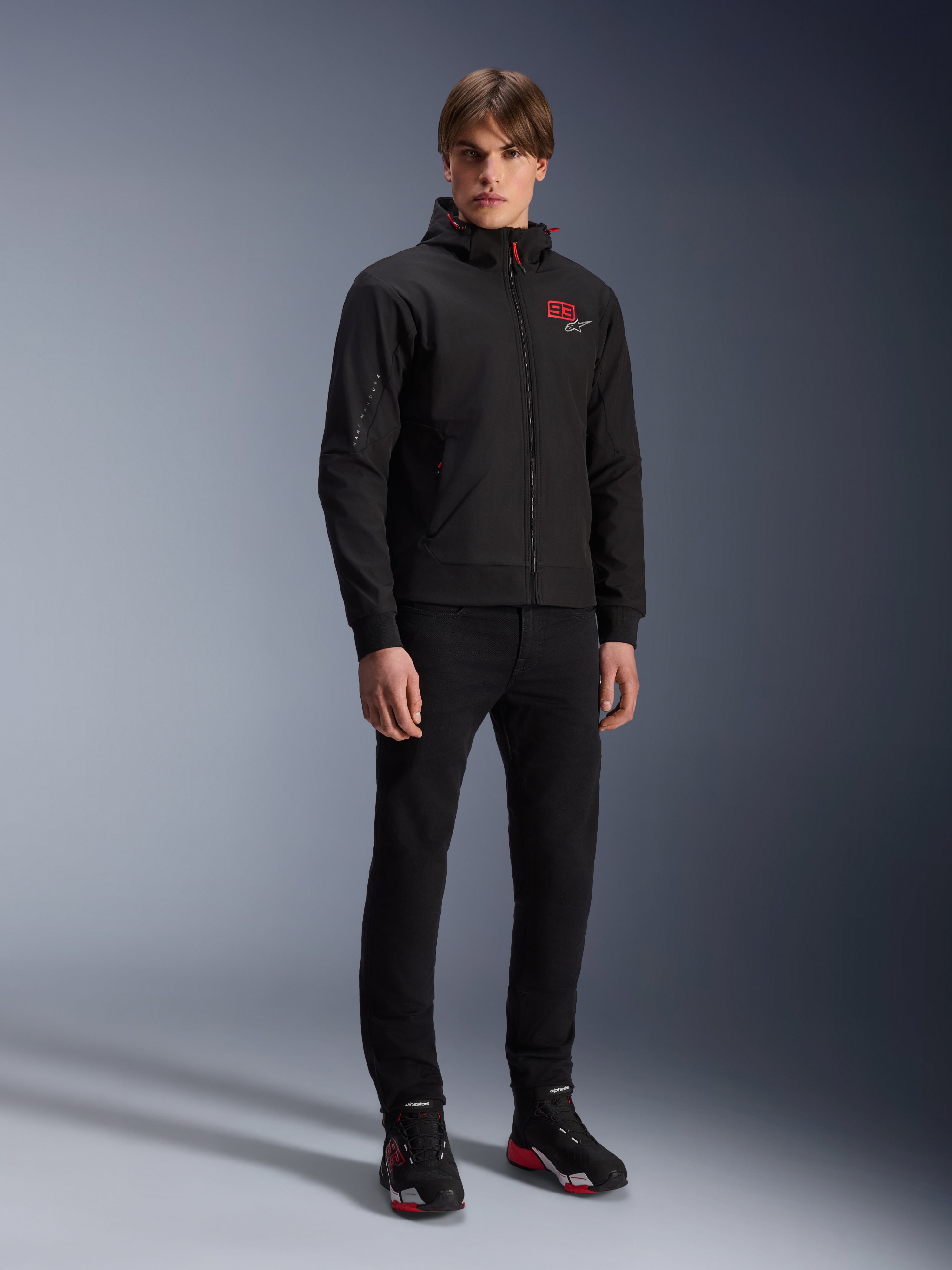 Sudadera con capucha Alpinestars MM93 Radium Tech, chaqueta de moto urbana, negra con detalles en rojo brillante, usada por un modelo con pantalones negros y calzado de moto, con el logotipo '93' rojo en el pecho y la marca Marc Márquez en la manga, ropa técnica casual para conducción urbana