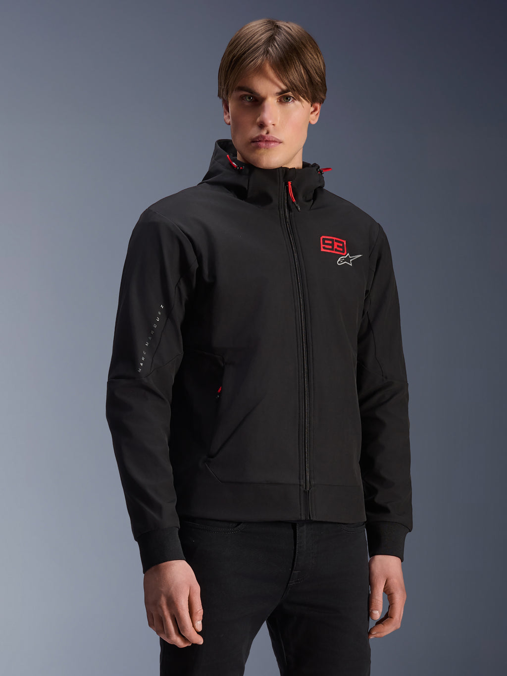 MM93 Radium Tech Hoodie, chaqueta de moto urbana negra con cremallera, número 93 en rojo y logo Alpinestars blanco en el pecho, lucida por un modelo con pantalones negros, con capucha extraíble con cordones rojos y marca en la manga, diseño casual inspirado en el motociclismo