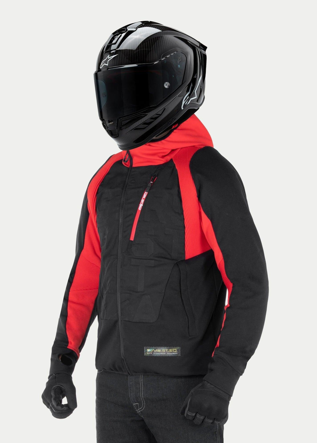 Chaqueta con capucha Alpinestars MO.ST.EQ Hybrid, sudadera urbana para moto en color negro y rojo, con cremallera frontal en ángulo y paneles acanalados en los hombros, combinada con un casco integral negro y guantes negros, estilo streetwear contemporáneo