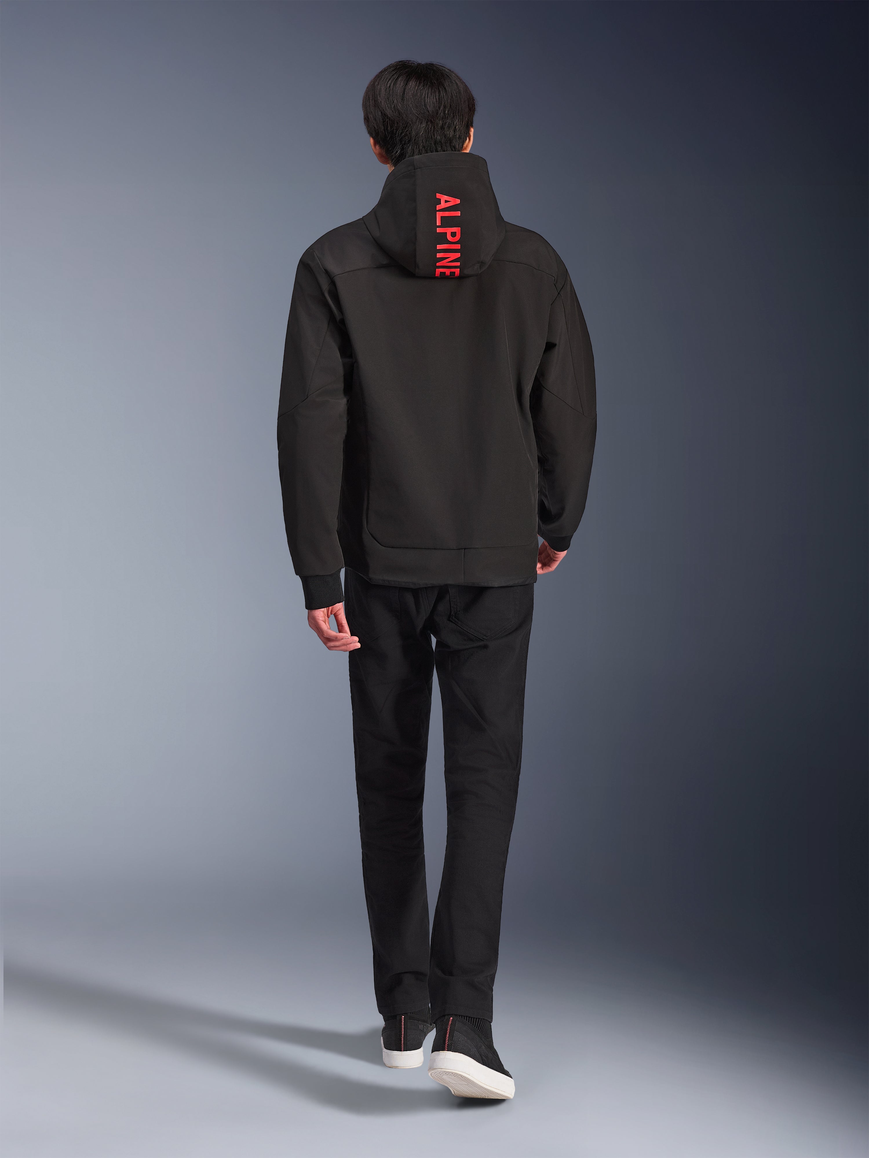 Sudadera con capucha Alpinestars Radium Tech, chaqueta de moto urbana, color negro, vista trasera con el texto 'ALPINE' en rojo brillante en la capucha, combinada con pantalones negros y zapatillas, elegante carcasa técnica para desplazamientos urbanos.
