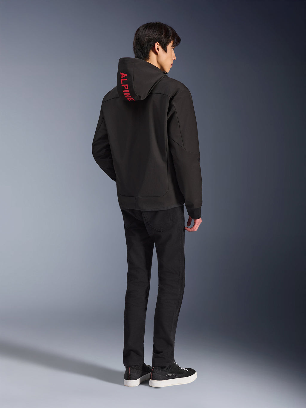 Alpinestars Radium Tech Hoodie, negra con la marca Alpinestars en rojo en la capucha, vista trasera en un modelo masculino con pantalones negros y zapatillas, equipamiento técnico para conducción urbana