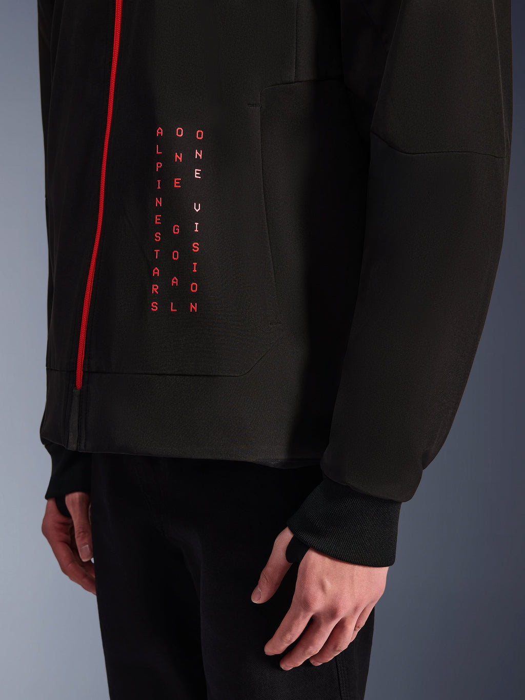 Alpinestars Radium Tech Hoodie, chaqueta de moto urbana, negro y rojo brillante, primer plano del gráfico frontal con texto vertical en rojo "ALPINESTARS ONE GOAL ONE VISION", cremallera roja y puño acanalado con orificio para el pulgar.