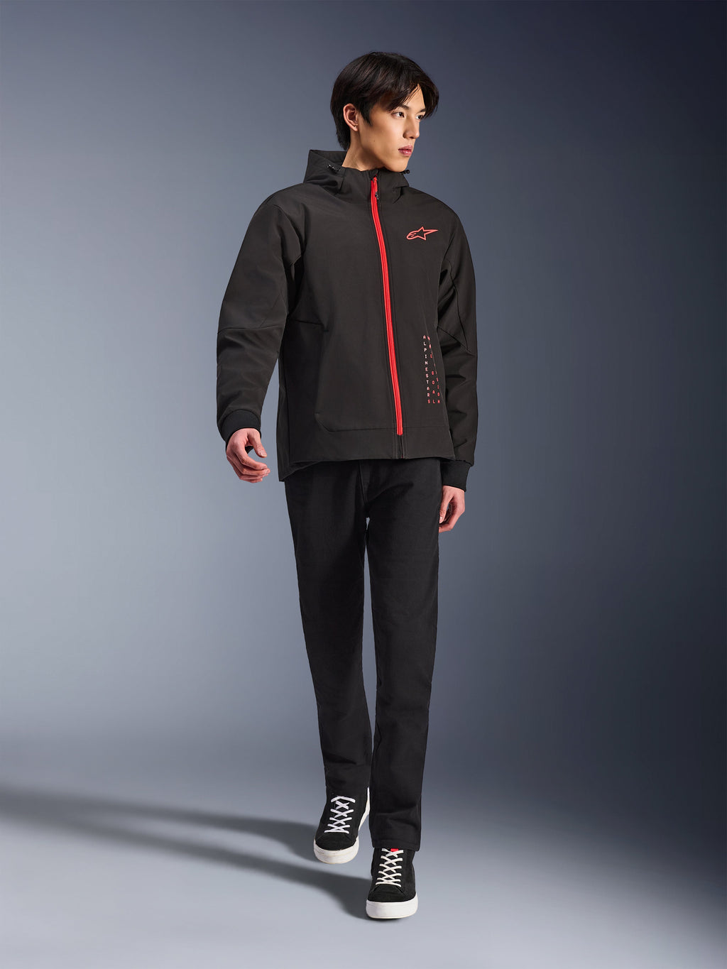 Alpinestars Radium Tech Hoodie en Black Bright Red, chaqueta de moto urbana usada por un modelo, con una cremallera frontal roja y el logo en el pecho, combinada con pantalones negros y zapatillas para un look moderno de trayecto diario.