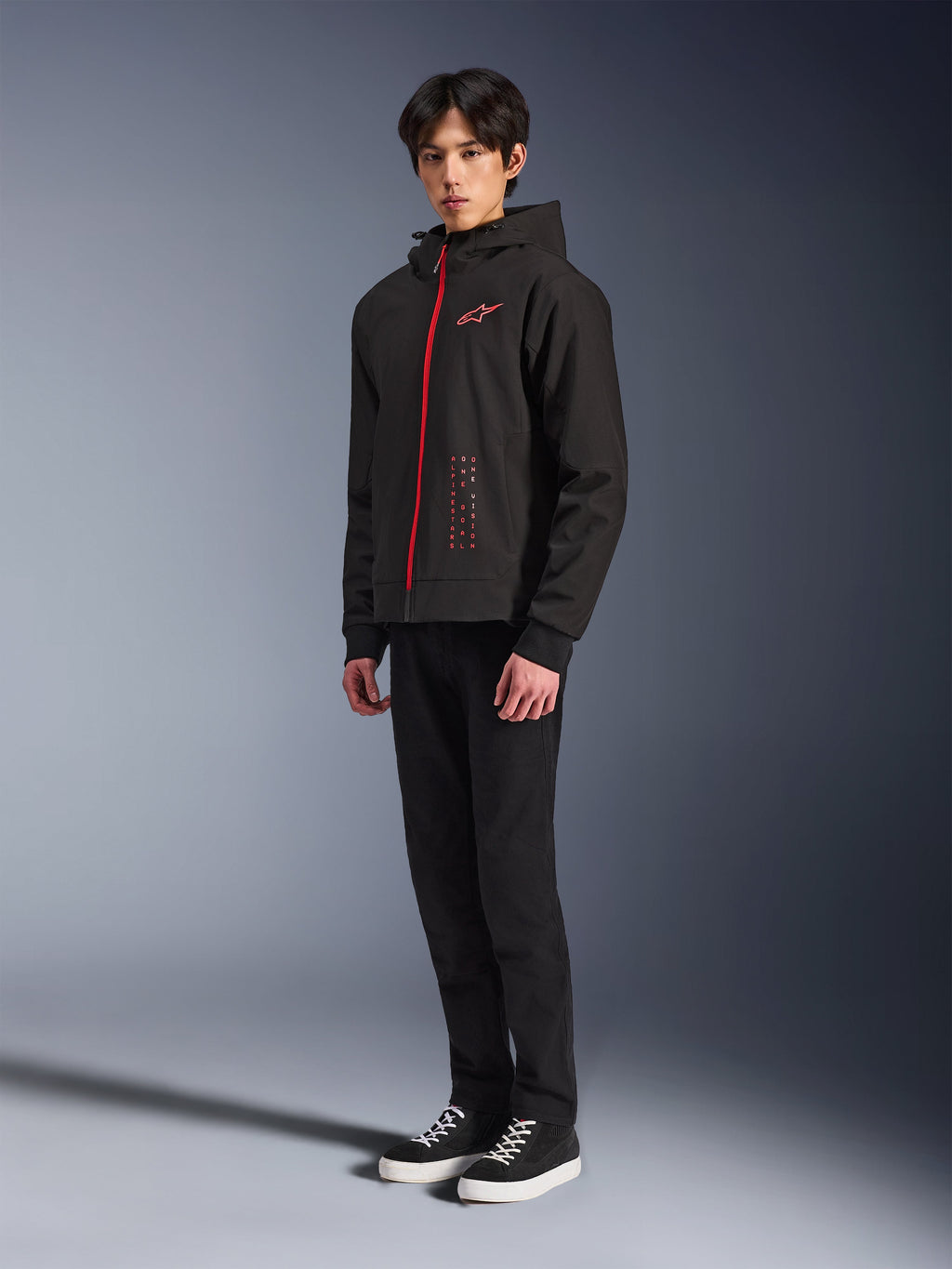 Alpinestars Radium Tech Hoodie, Black Bright Red, sudadera técnica para conducción urbana con cremallera frontal roja y logotipo en el pecho, vestida por un modelo con pantalones negros y zapatillas negras con suelas blancas, toma de estudio de cuerpo completo.