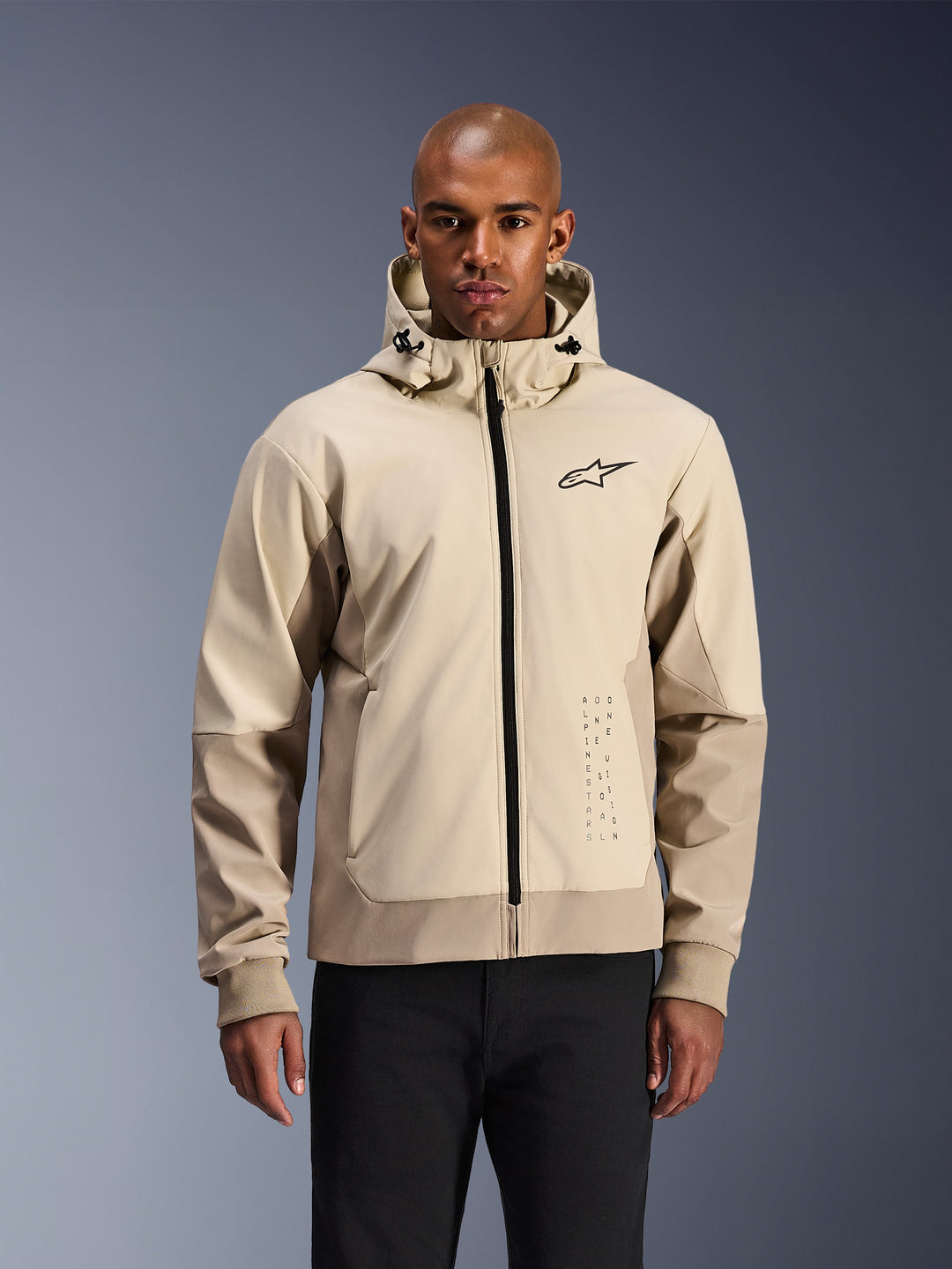 Alpinestars Radium Tech Hoodie en Mastic Aluminum, chaqueta de moto urbana en beige claro y tostado, cremallera frontal negra y logotipo pequeño en el pecho, modelada por un hombre con pantalones negros, exterior técnico cortavientos con estilo streetwear contemporáneo
