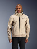 Alpinestars Radium Tech Hoodie en Mastic Aluminum, chaqueta de moto urbana en beige claro y tostado, cremallera frontal negra y logotipo pequeño en el pecho, modelada por un hombre con pantalones negros, exterior técnico cortavientos con estilo streetwear contemporáneo
