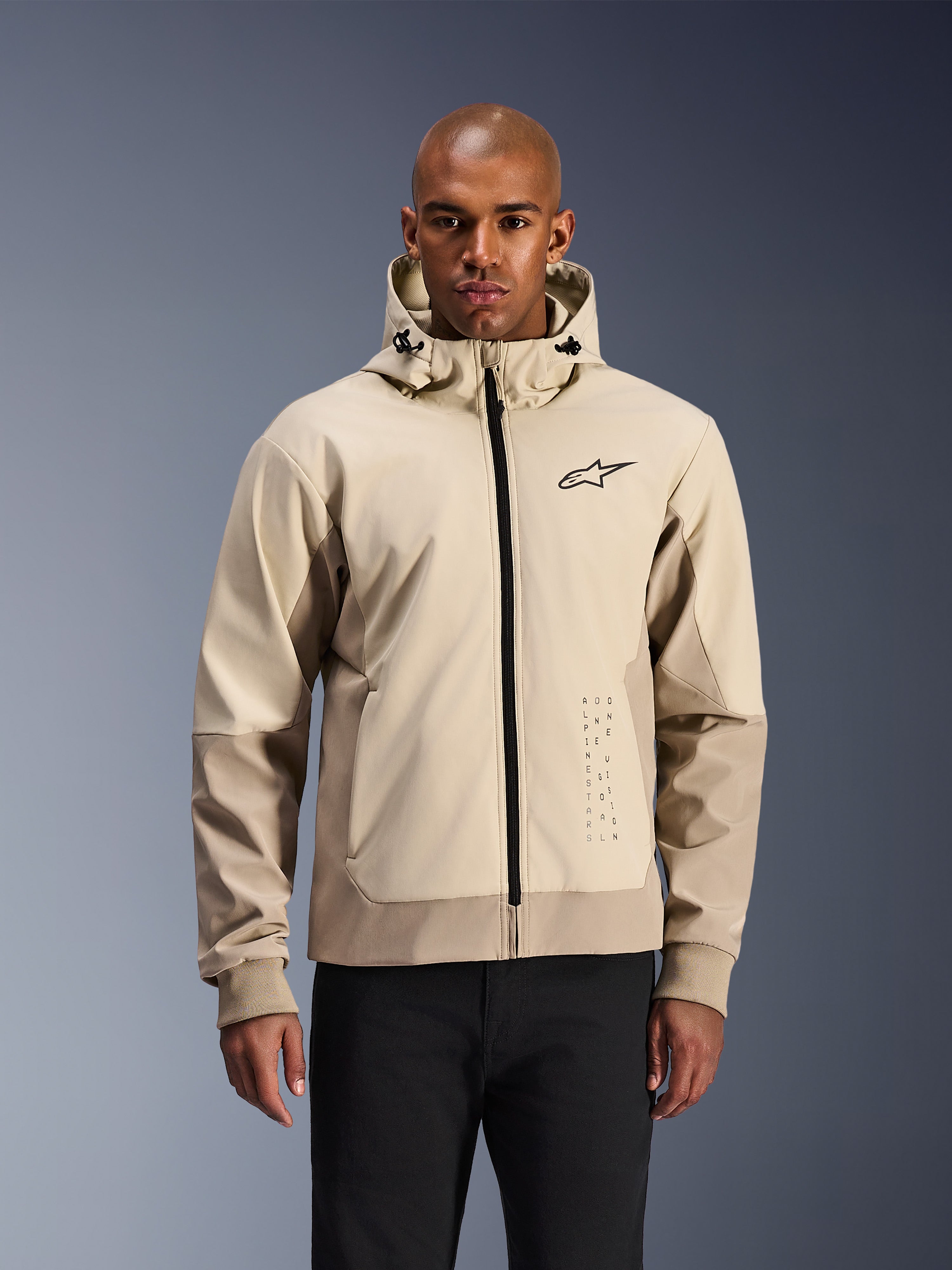 Alpinestars Radium Tech Hoodie en Mastic Aluminum, chaqueta de moto urbana en beige claro y tostado, cremallera frontal negra y logotipo pequeño en el pecho, modelada por un hombre con pantalones negros, exterior técnico cortavientos con estilo streetwear contemporáneo