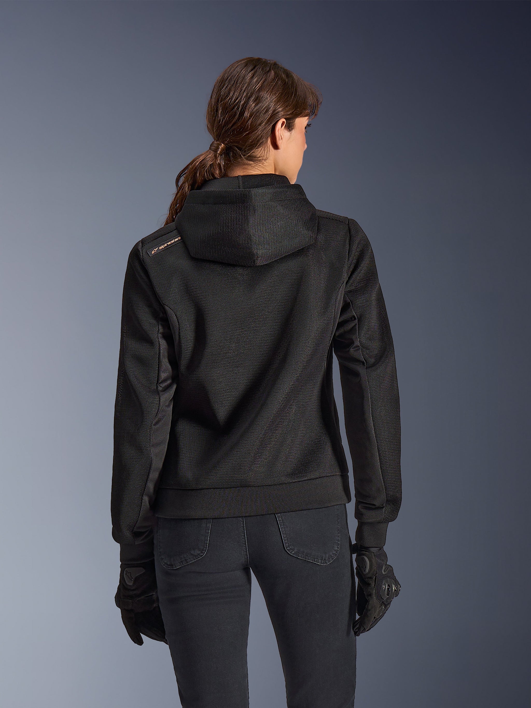 Alpinestars Stella Chrome Superairflow Hoodie, vista trasera de mujer con sudadera técnica de malla 3D para moto en color negro, cuenta con capucha técnica y logotipo de color cobre en el hombro, usada con guantes y pantalones de motociclismo negros.