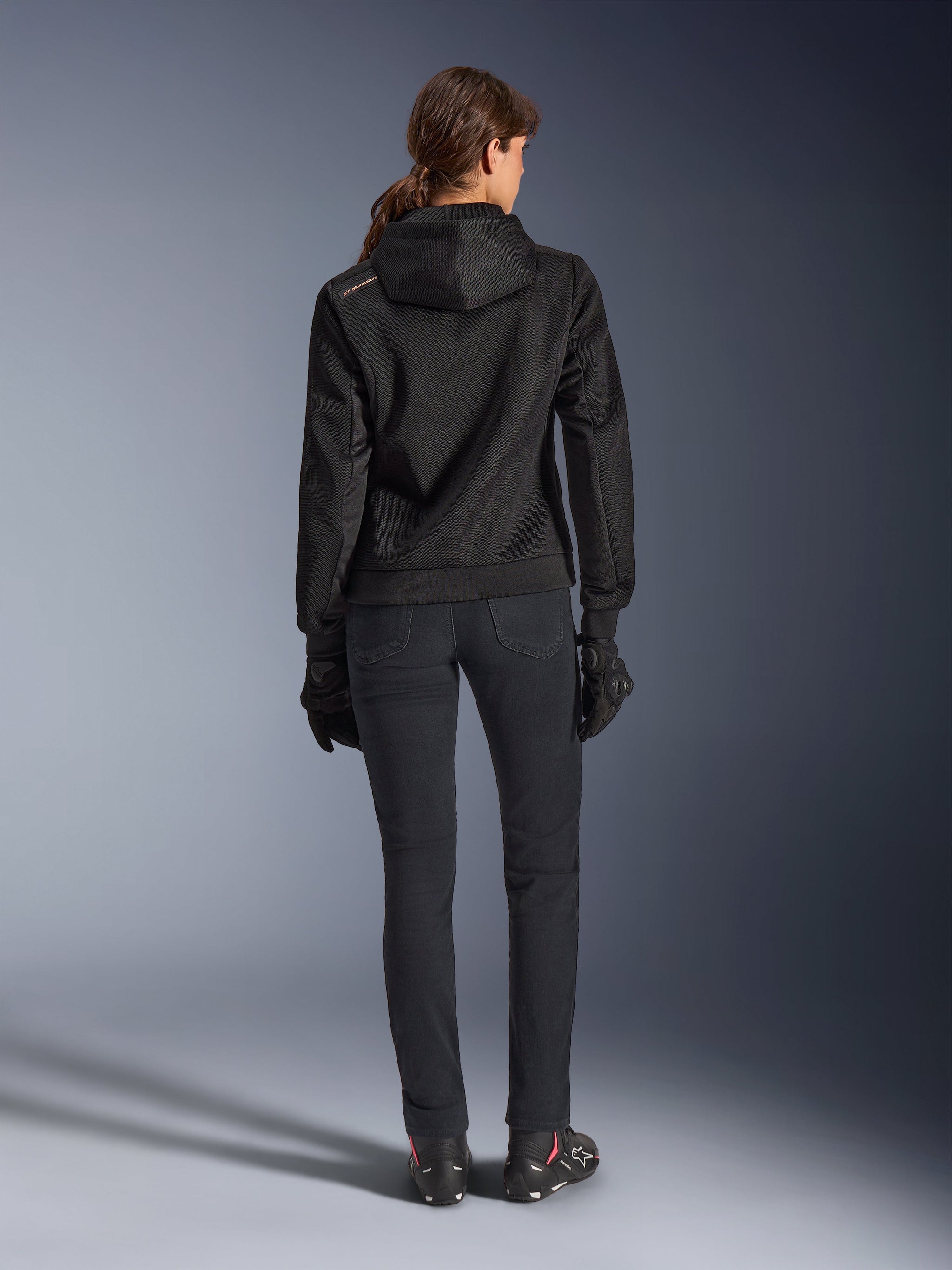 Alpinestars Stella Chrome Superairflow Hoodie, sudadera técnica de moto para mujer, malla 3D negra con pequeño logotipo naranja en el hombro, vista trasera, combinada con pantalones de conducción negros, guantes y botas negros, diseñada para conducción urbana deportiva