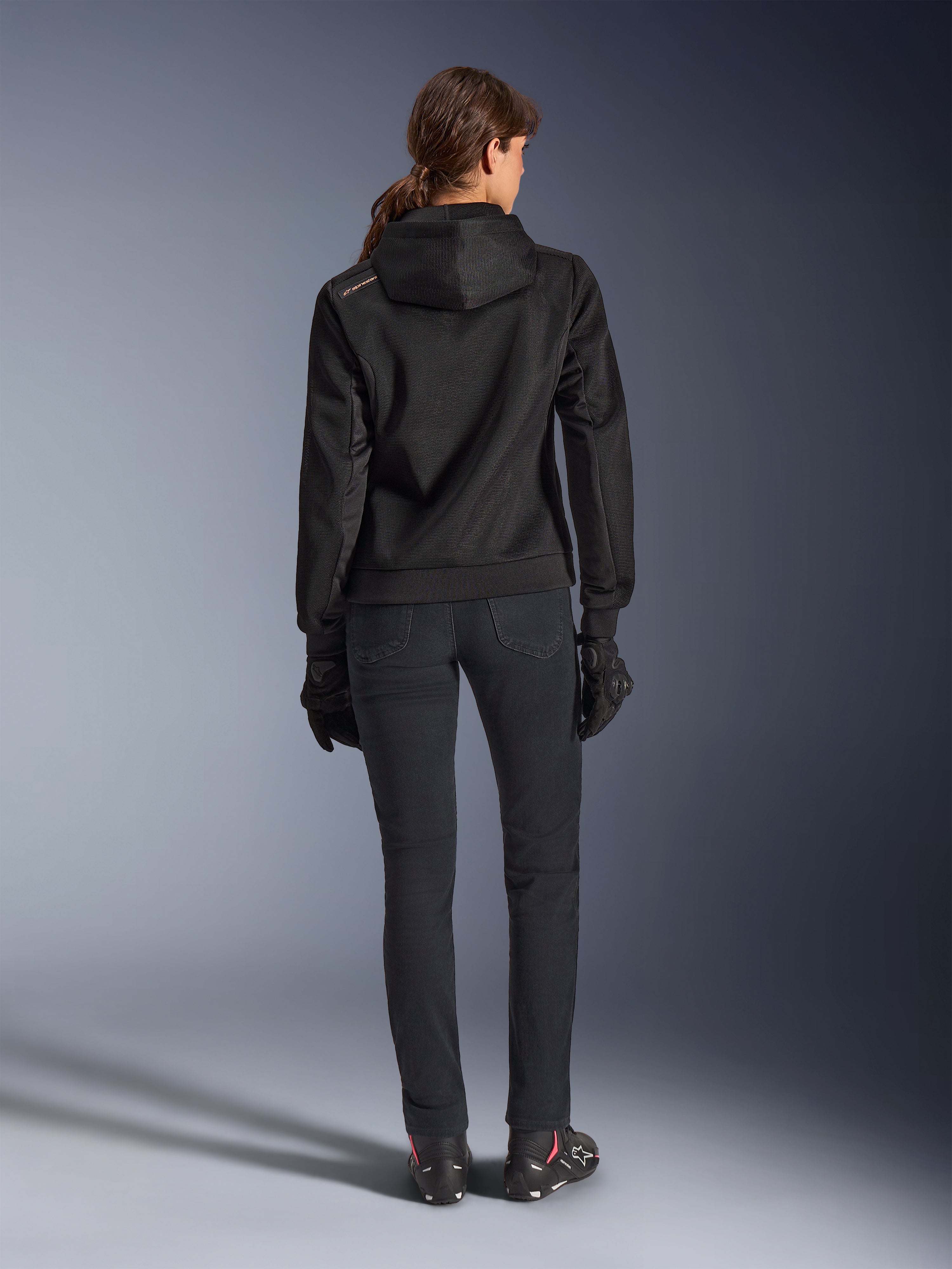 Alpinestars Stella Chrome Superairflow Hoodie, sudadera técnica de moto para mujer, malla 3D negra con pequeño logotipo naranja en el hombro, vista trasera, combinada con pantalones de conducción negros, guantes y botas negros, diseñada para conducción urbana deportiva
