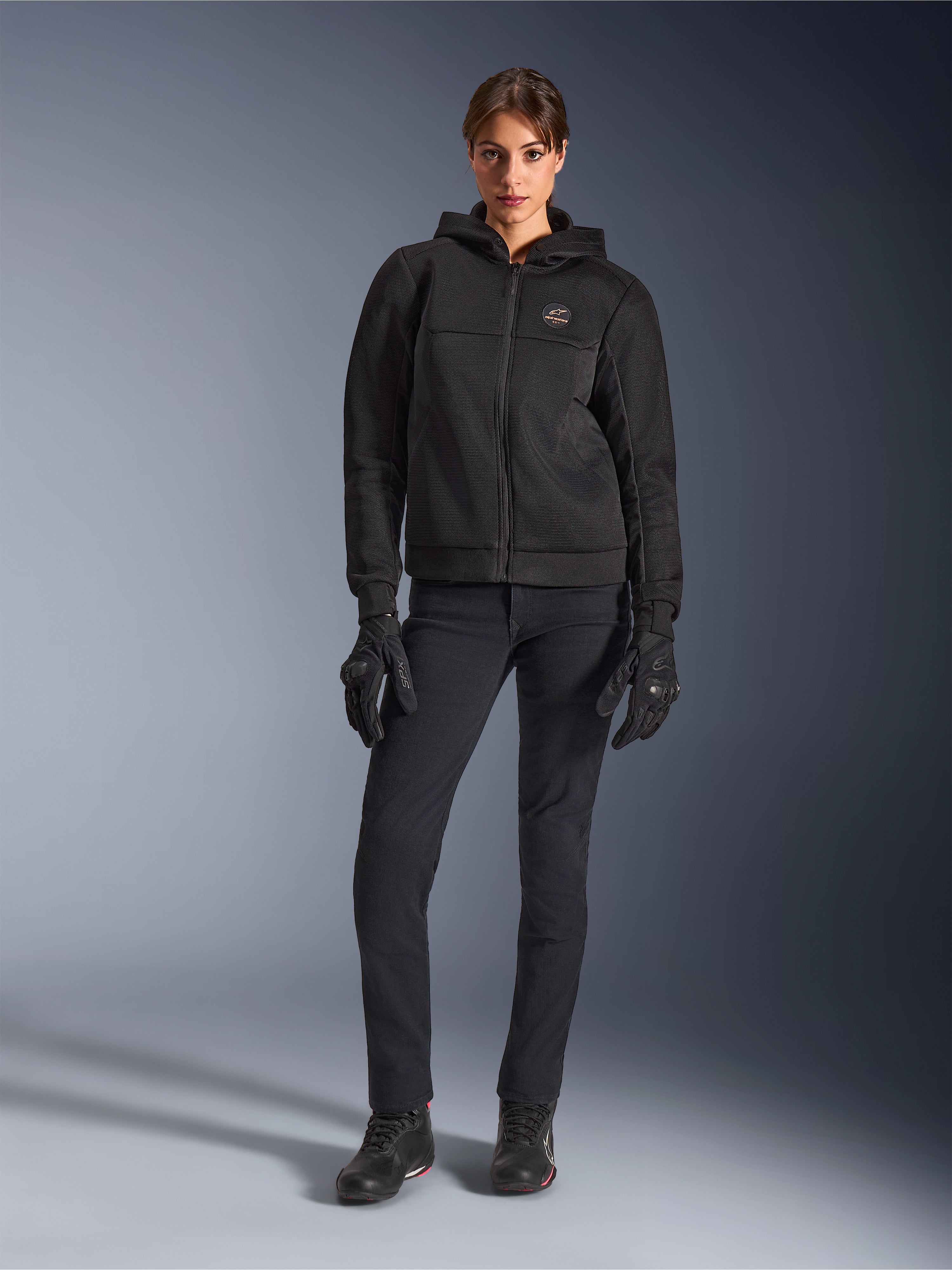 Modelo femenina luciendo la sudadera Alpinestars Stella Chrome Superairflow en color negro, combinada con pantalones técnicos de conducción y guantes negros, con construcción de malla 3D y logotipo en el pecho en tono cobre, ropa de motociclismo deportiva urbana para mujer.
