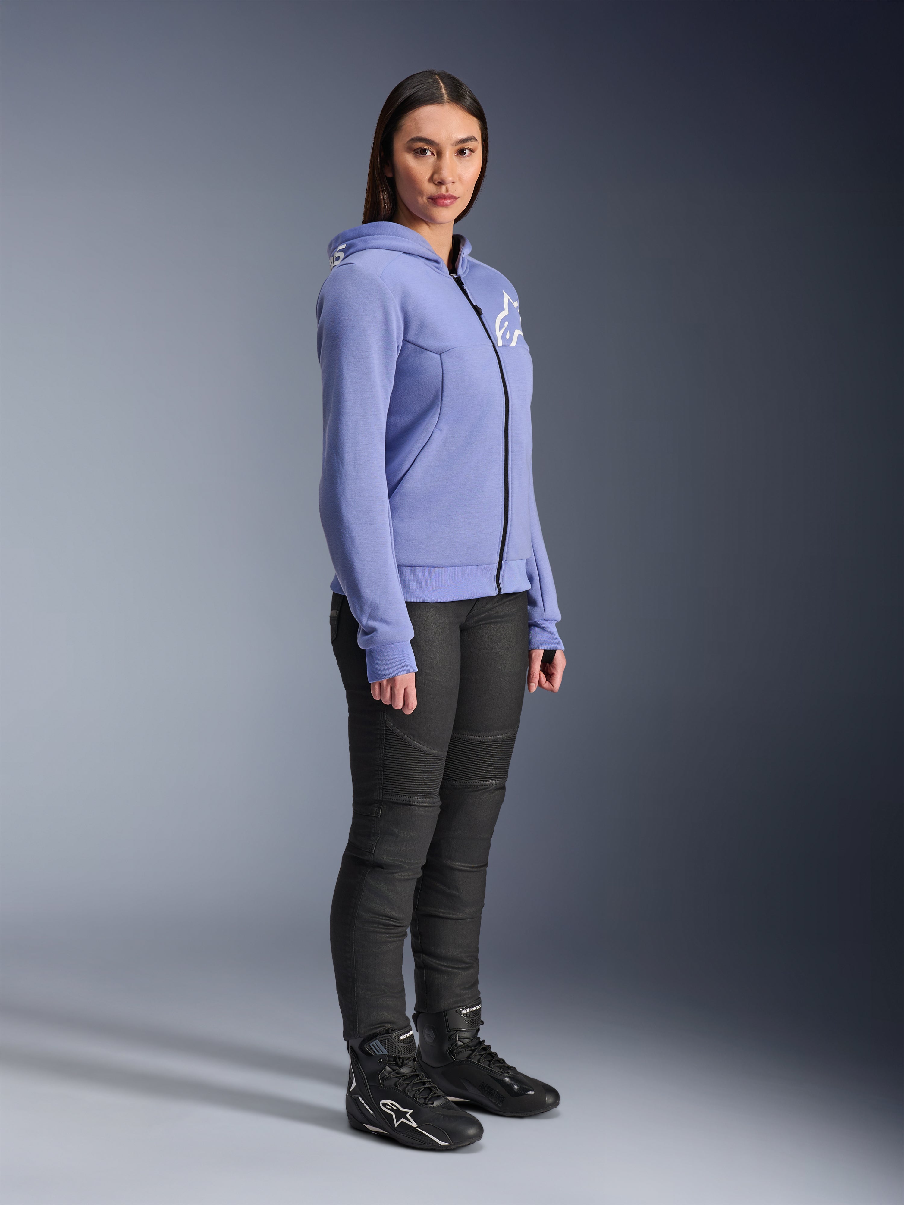 Alpinestars Woman Stella Chrome V2 Sport Hoodie, sudadera de moto en color azul violeta con el logotipo blanco de Alpinestars en el pecho, vestida por una modelo con pantalones de montar negros y botas de moto negras, ropa de conducción urbana para mujer