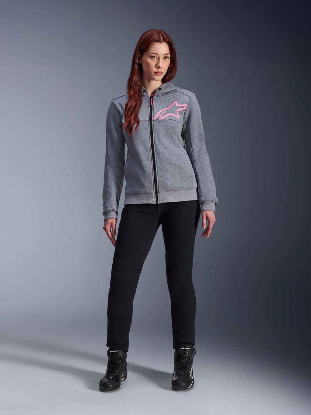 Alpinestars Stella Chrome V2 Sport Hoodie, gris jaspeado rosa flúor, vestida por una mujer con pantalones negros y botas, con el logotipo de la estrella Alpinestars en rosa en el pecho, equipamiento urbano para moto con cremallera completa, forro polar técnico ligero con un ajuste atlético casual.