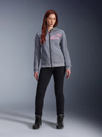 Alpinestars Stella Chrome V2 Sport Hoodie, gris jaspeado rosa flúor, vestida por una mujer con pantalones negros y botas, con el logotipo de la estrella Alpinestars en rosa en el pecho, equipamiento urbano para moto con cremallera completa, forro polar técnico ligero con un ajuste atlético casual.
