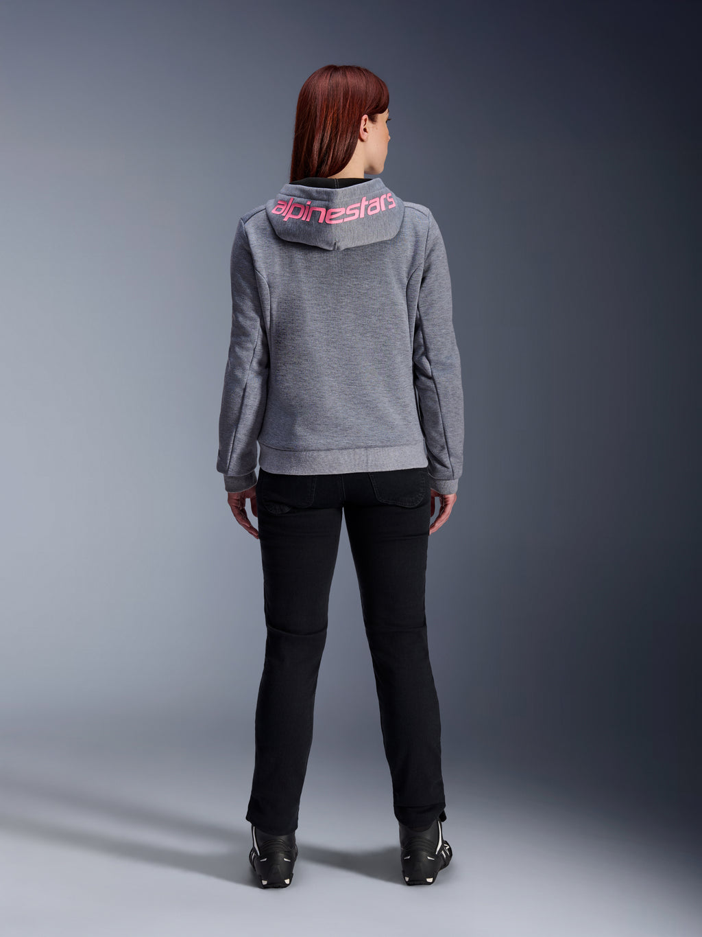 Alpinestars Stella Chrome V2 Sport Hoodie, sudadera de moto para mujer, gris jaspeado y rosa flúor, vista posterior que muestra el logotipo rosa de Alpinestars en la capucha, combinada con pantalones de conducción negros y botas, ropa de moto casual para mujer