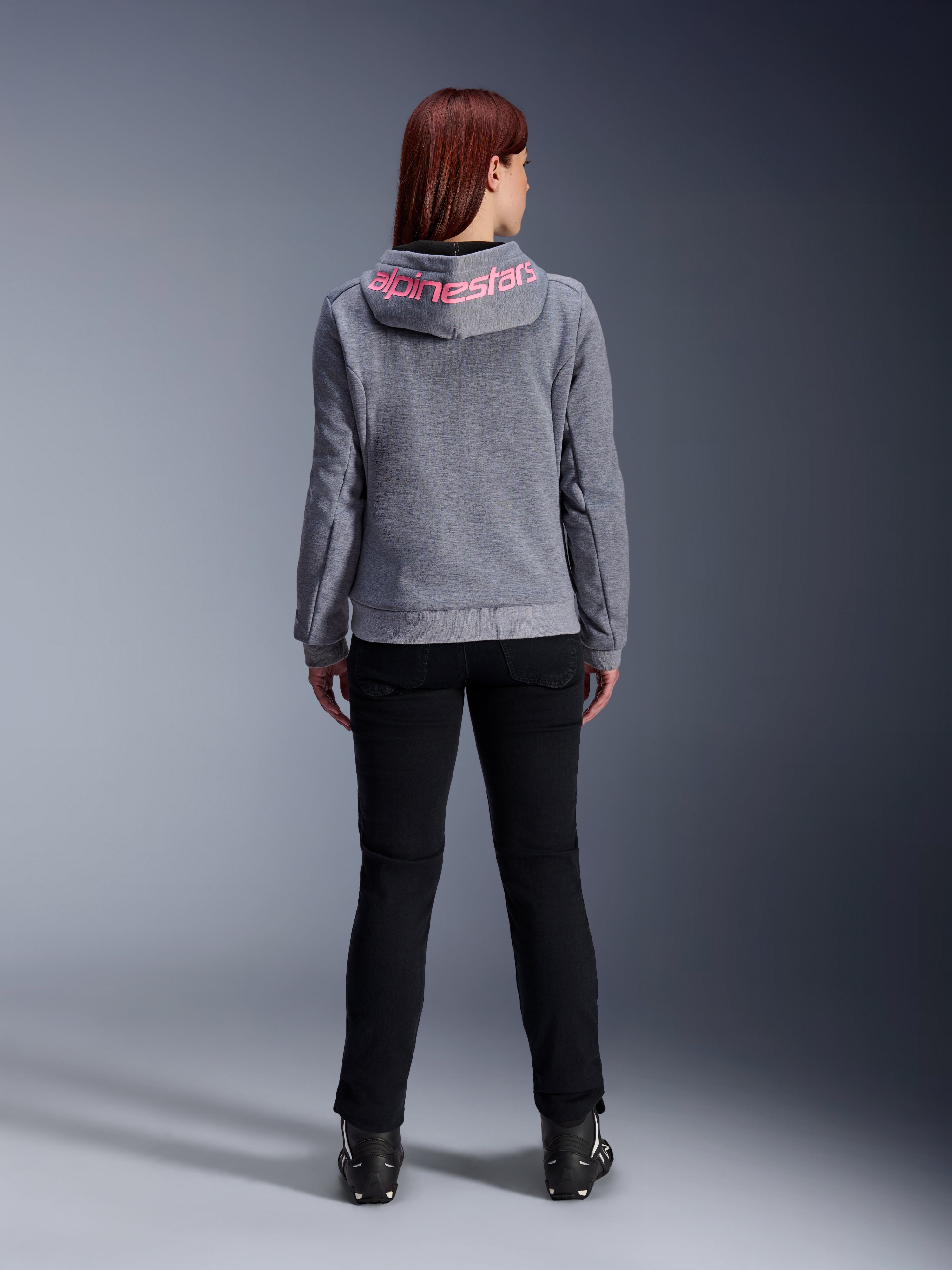 Alpinestars Stella Chrome V2 Sport Hoodie, sudadera de moto para mujer, gris jaspeado y rosa flúor, vista posterior que muestra el logotipo rosa de Alpinestars en la capucha, combinada con pantalones de conducción negros y botas, ropa de moto casual para mujer