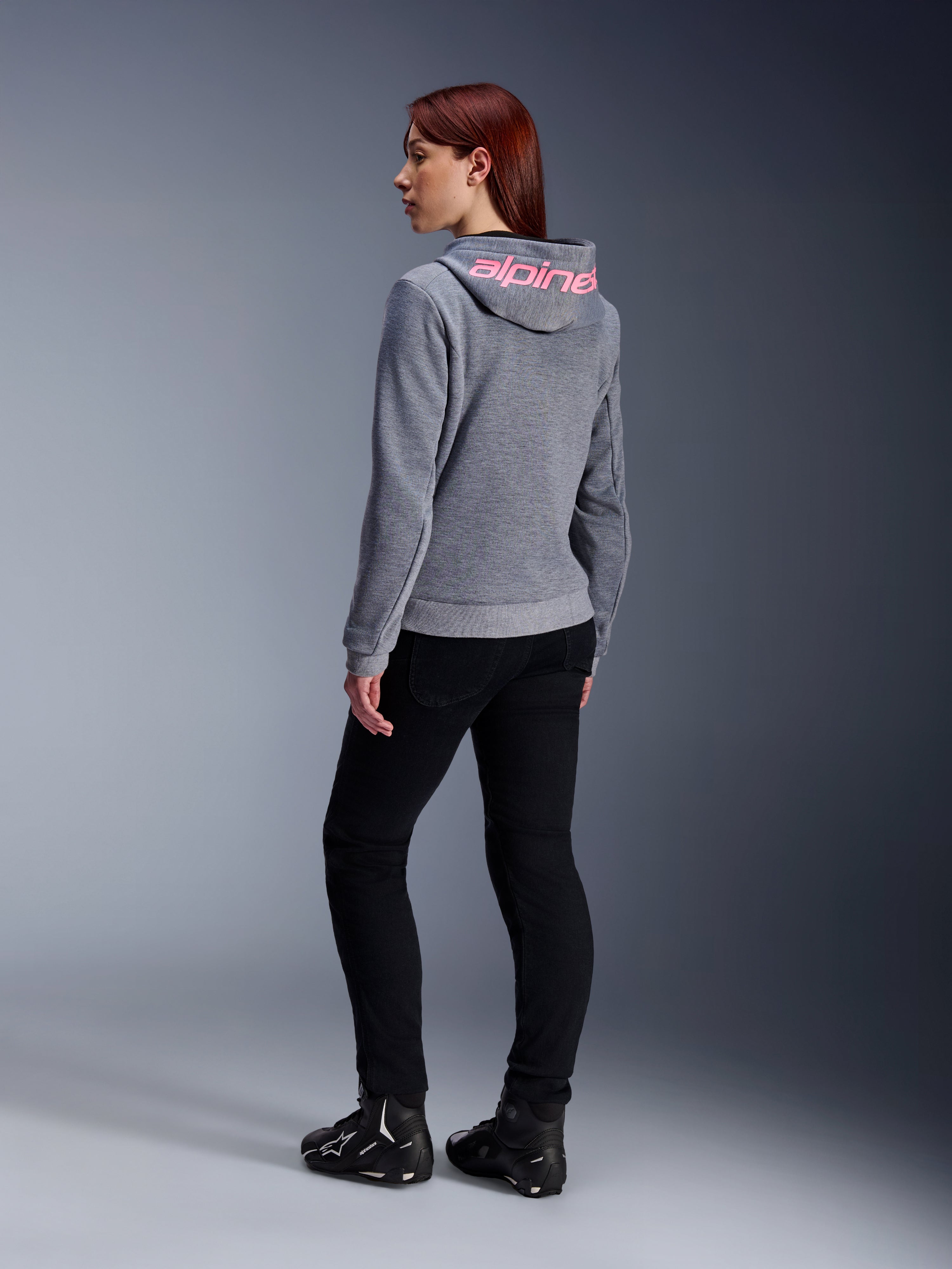 Alpinestars Stella Chrome V2 Sport Hoodie, sudadera de moto para mujer, gris jaspeado con logotipo rosa flúor en la capucha, vista trasera, combinada con pantalones negros y calzado de moto, estilo urbano casual con protección para motociclistas