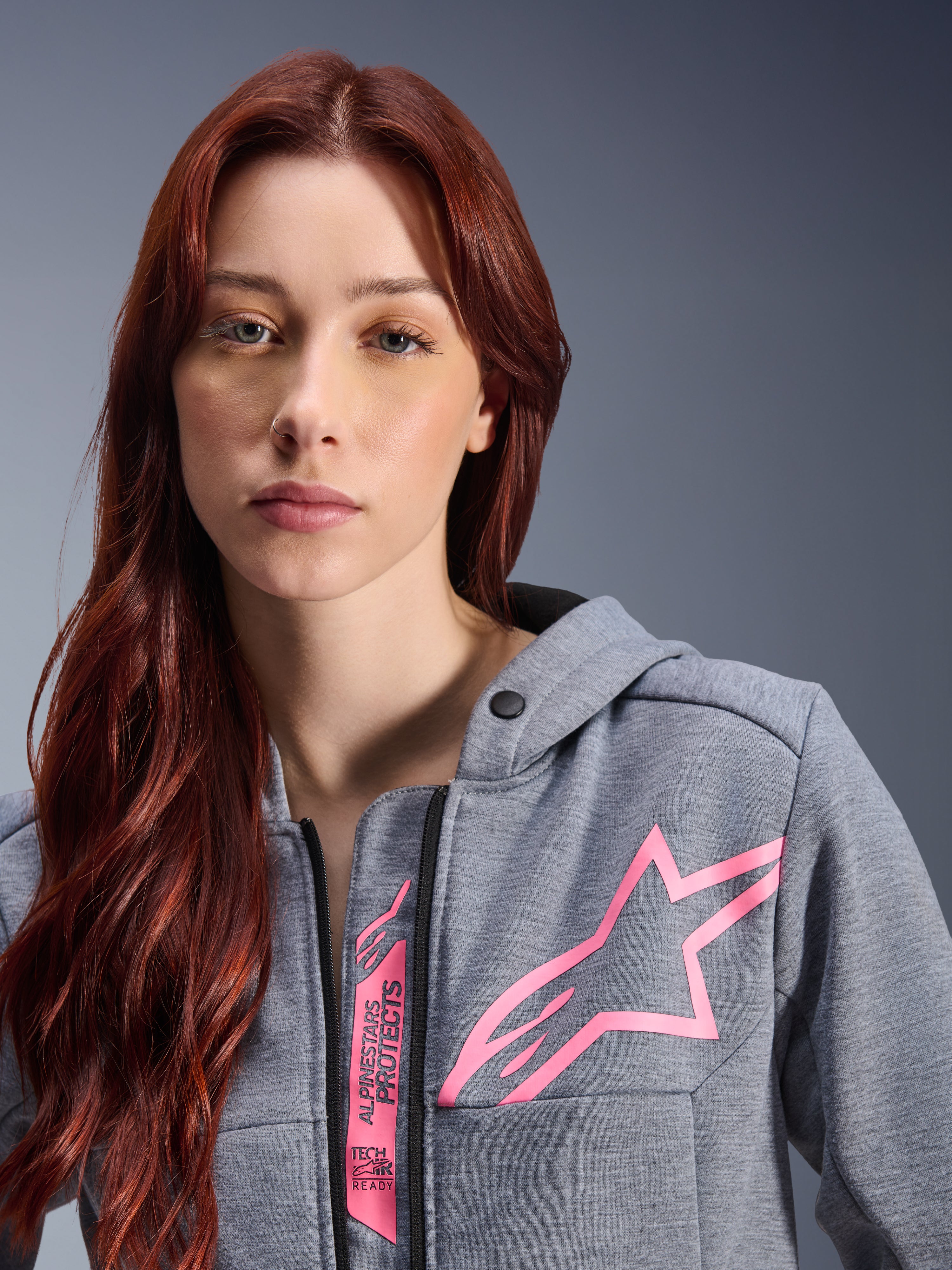 Mujer con la sudadera Alpinestars Stella Chrome V2 Sport Hoodie, gris jaspeado y rosa flúor, primer plano que muestra el logotipo de la estrella rosa de Alpinestars y la marca Tech-Air Ready, ropa urbana para moto