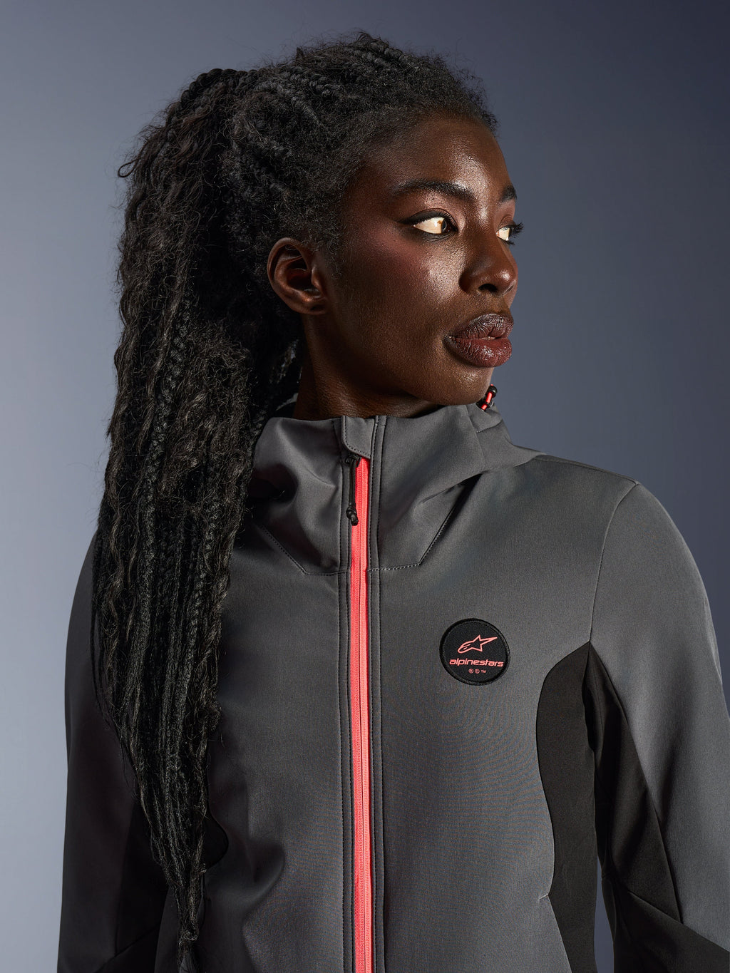 Modelo femenina luciendo la sudadera Alpinestars Stella Radium Tech Hoodie en gris intenso y negro, con una cremallera en color coral brillante, logotipo circular en el pecho y capa exterior cortavientos de cuello alto, equipamiento urbano informal de motociclismo para mujer