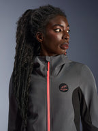 Modelo femenina luciendo la sudadera Alpinestars Stella Radium Tech Hoodie en gris intenso y negro, con una cremallera en color coral brillante, logotipo circular en el pecho y capa exterior cortavientos de cuello alto, equipamiento urbano informal de motociclismo para mujer