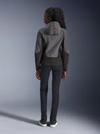 Mujer con sudadera técnica Alpinestars Stella Radium, gris oscuro y negro, vista trasera que muestra la marca 'ALPIN' en la capucha, combinada con pantalones negros de corte ajustado y zapatillas negras, ropa urbana para motociclismo