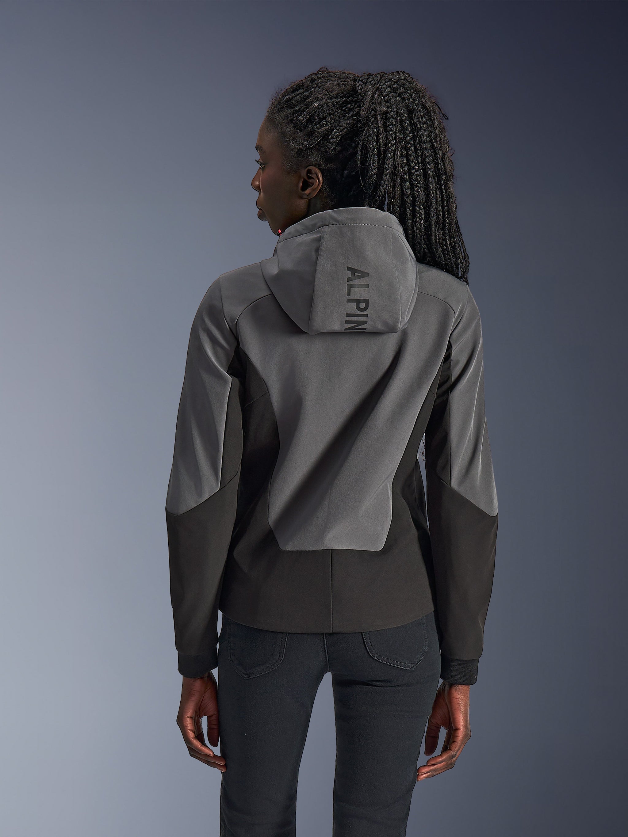 Woman wearing Alpinestars Stella Radium Tech Hoodie, rear view, Deep Gray Black, two-tone gray and black 3-layer windproof shell, Alpinestars branding on the hood, athletic fit urban motorcycle apparel