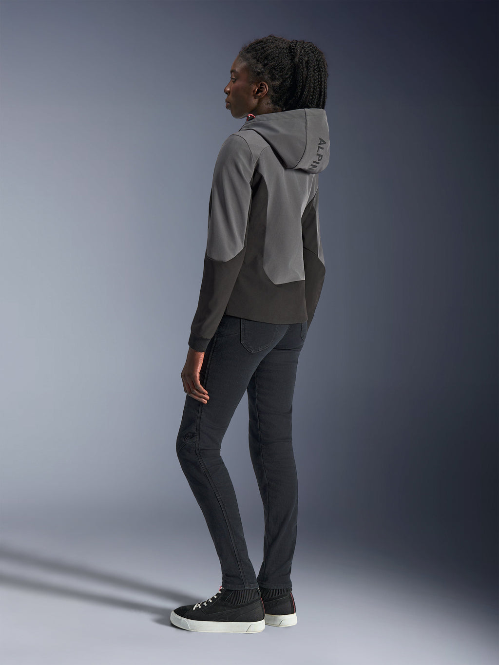 Mujer vistiendo la sudadera Alpinestars Stella Radium Tech en Gris Oscuro y Negro, ropa urbana para motociclismo, vista posterior que muestra la capucha con la marca Alpinestars, combinada con pantalones negros de corte ajustado y zapatillas de caña alta