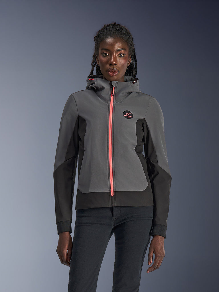 Alpinestars Stella Radium Tech Hoodie, sudadera de moto para mujer, gris oscuro y negro con cremallera y cordones de la capucha en rosa vibrante, usada por una modelo, diseño urbano deportivo con logotipo de Alpinestars en el pecho