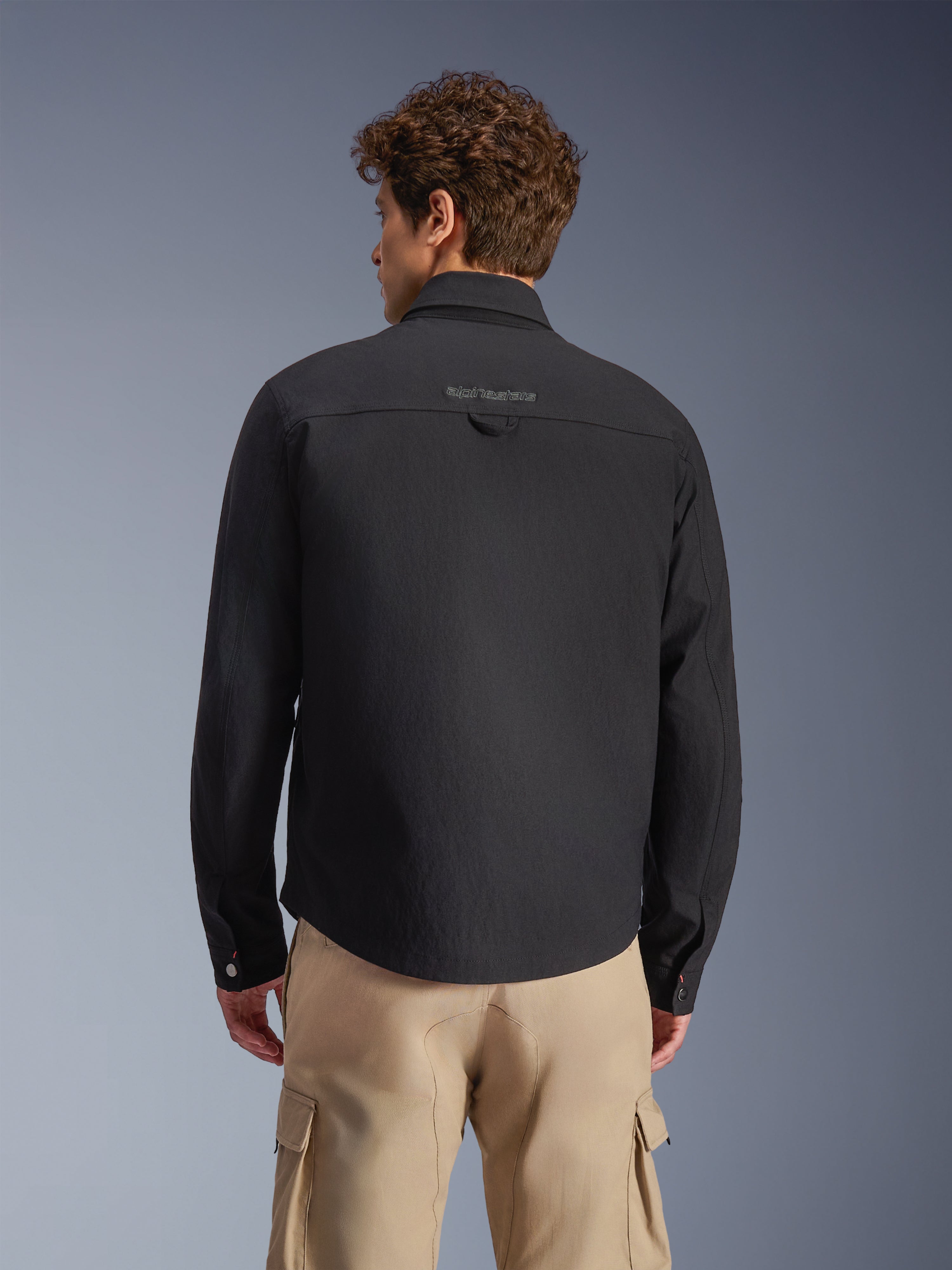 Alpinestars Bryon WR Sobrecamisa, negra, vista trasera llevada por un modelo, con marca bordada en el canesú trasero y trabilla de utilidad, combinada con pantalones cargo color canela, equipamiento urbano para moto con estética clásica de ropa de trabajo