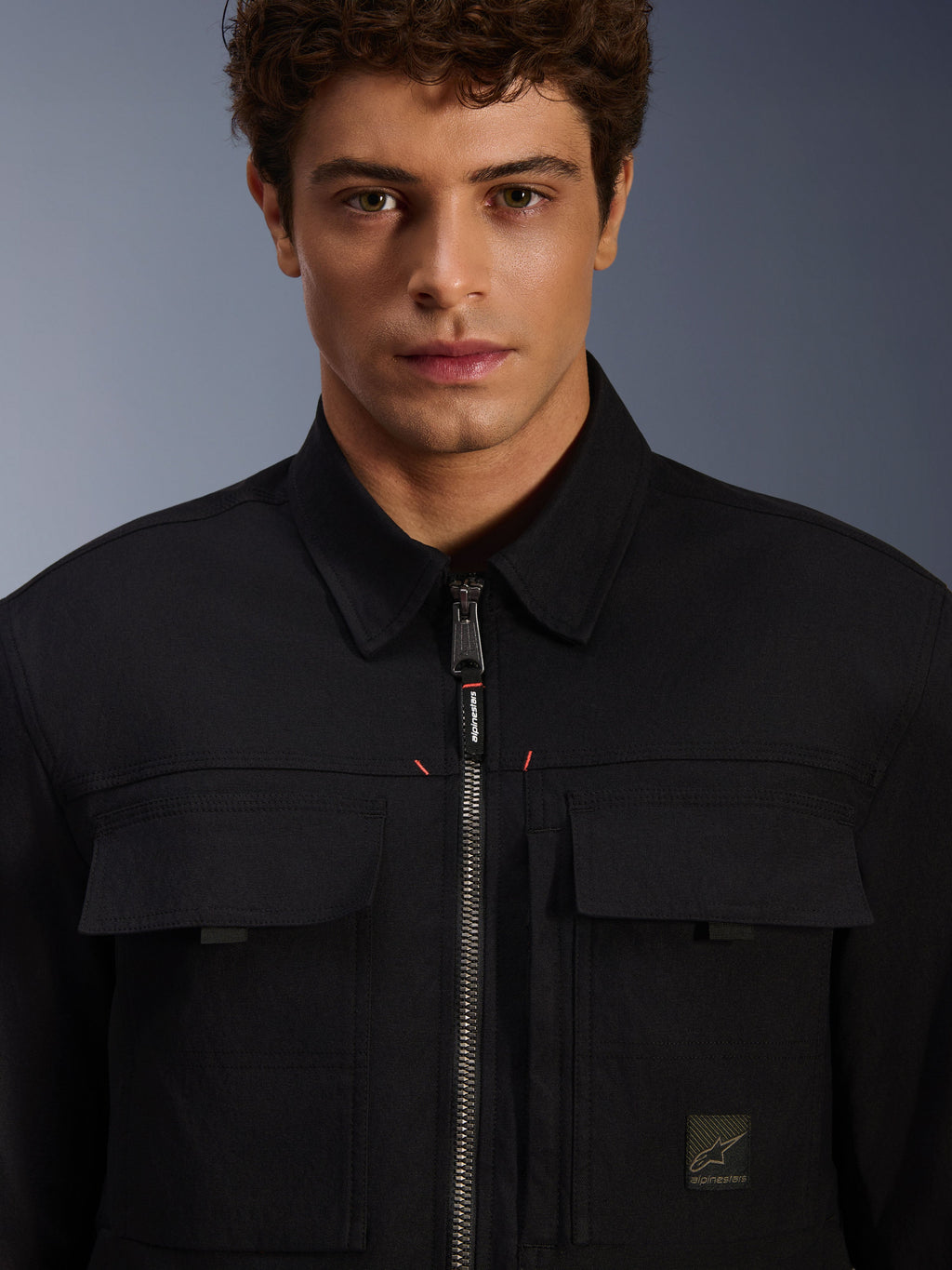 Alpinestars Bryon Water Resistant Overshirt, negra, vista de primer plano de la cremallera frontal y los bolsillos del pecho, textura de lona encerada duradera, diseño urbano minimalista con detalles de la marca y etiqueta de logotipo tejida