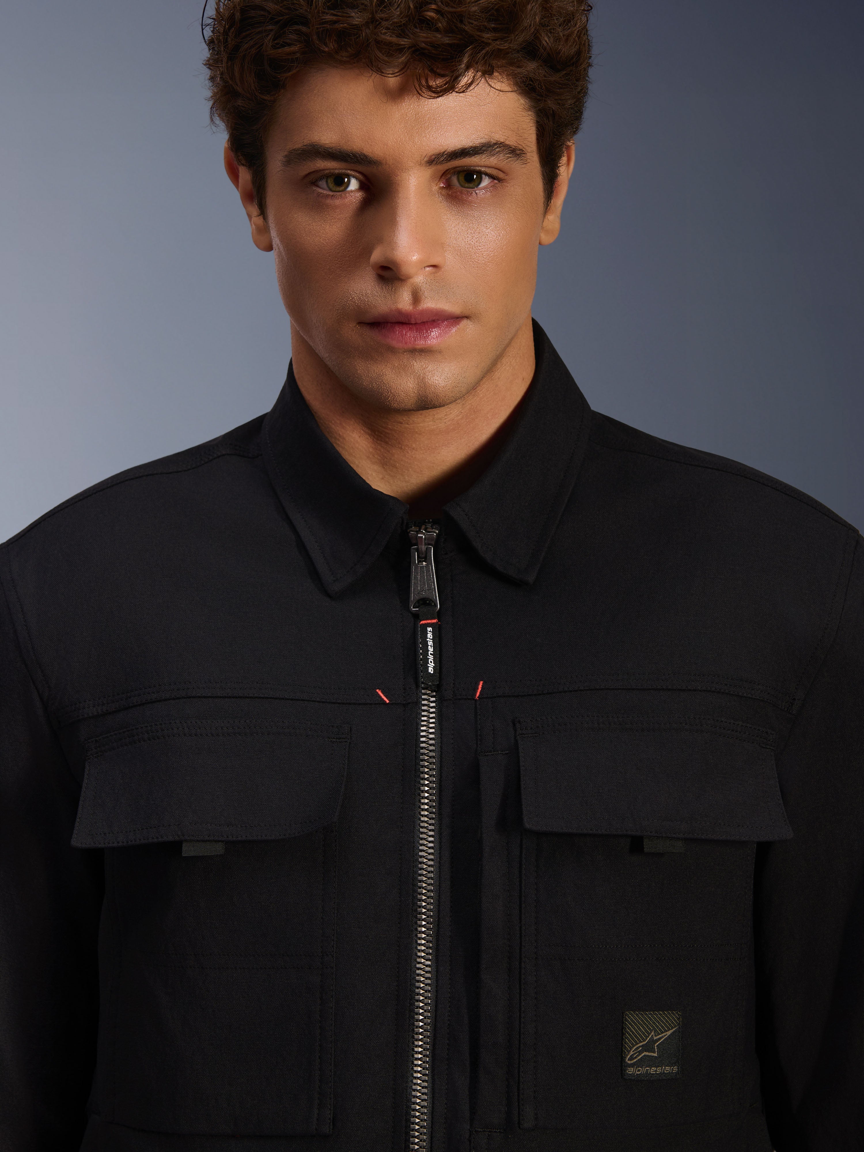 Alpinestars Bryon Water Resistant Overshirt, negra, vista de primer plano de la cremallera frontal y los bolsillos del pecho, textura de lona encerada duradera, diseño urbano minimalista con detalles de la marca y etiqueta de logotipo tejida