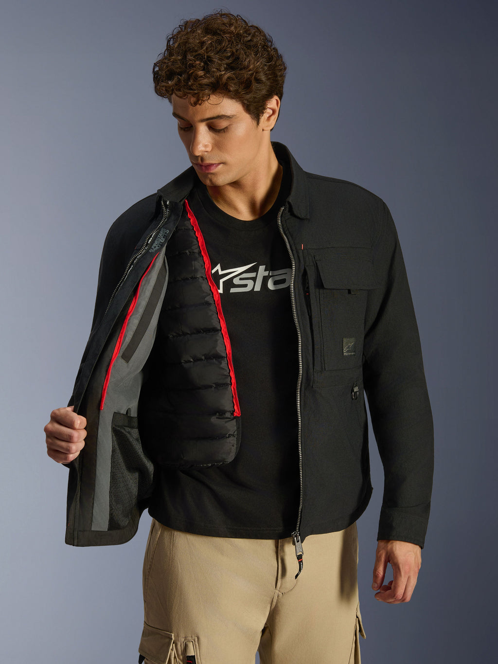 Sobrecamisa Alpinestars Bryon WR, negra, chaqueta de moto urbana mostrada abierta para revelar un chaleco térmico acolchado negro desmontable con ribete rojo, combinada con pantalones cargo tostados y camiseta con logotipo, diseño funcional inspirado en la ropa de trabajo
