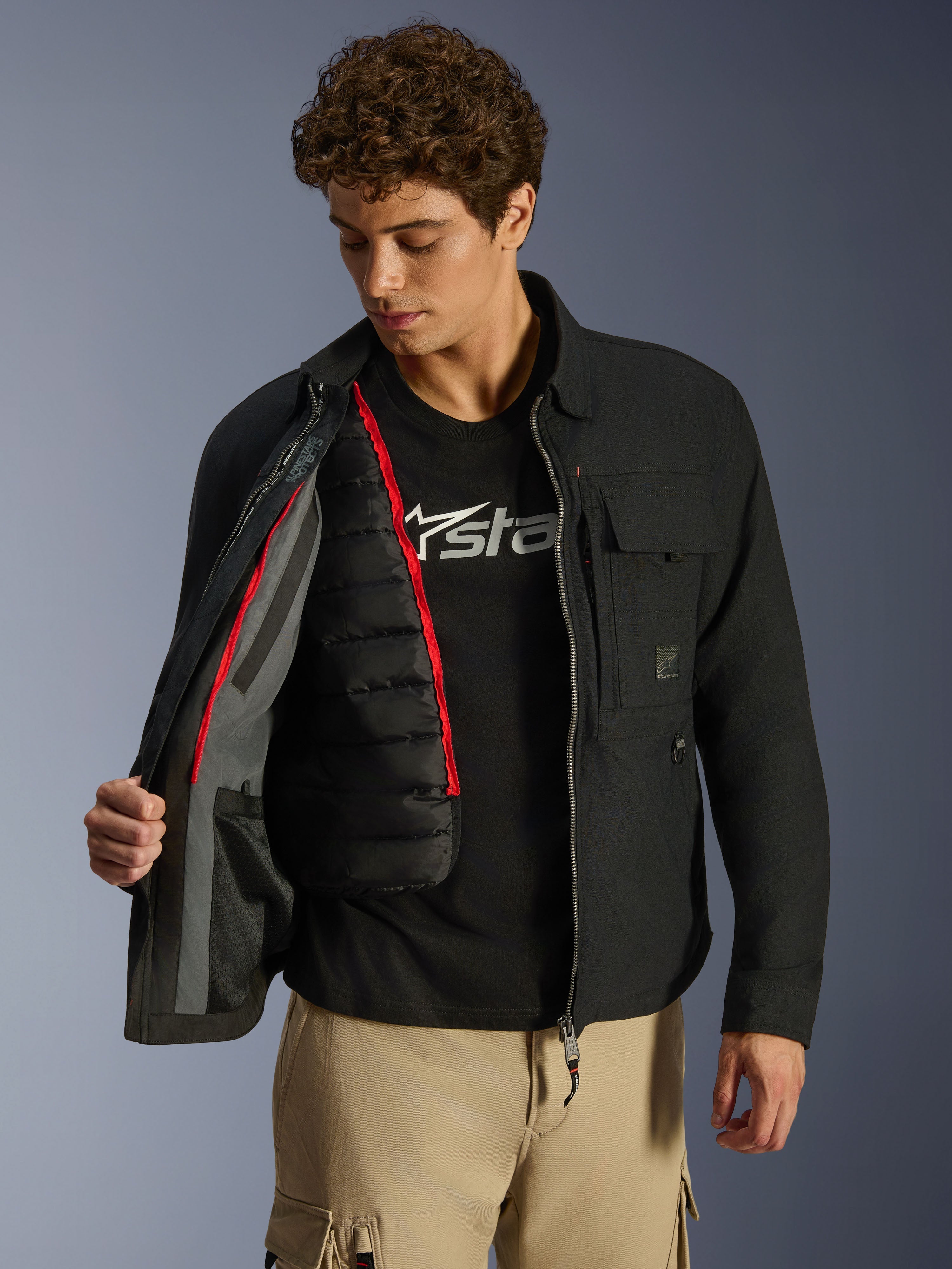 Sobrecamisa Alpinestars Bryon WR, negra, chaqueta de moto urbana mostrada abierta para revelar un chaleco térmico acolchado negro desmontable con ribete rojo, combinada con pantalones cargo tostados y camiseta con logotipo, diseño funcional inspirado en la ropa de trabajo