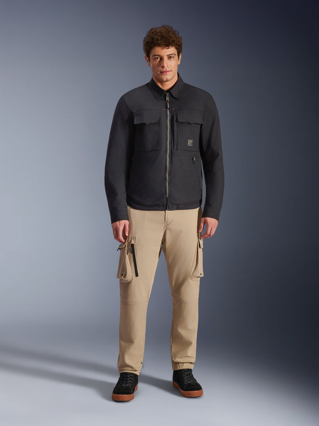 Modelo masculino vistiendo la sobrecamisa Alpinestars Bryon Water Resistant en color negro, con bolsillos cargo en el pecho y cremallera central, combinada con pantalones cargo color canela y zapatillas negras, retrato de estudio de cuerpo completo.
