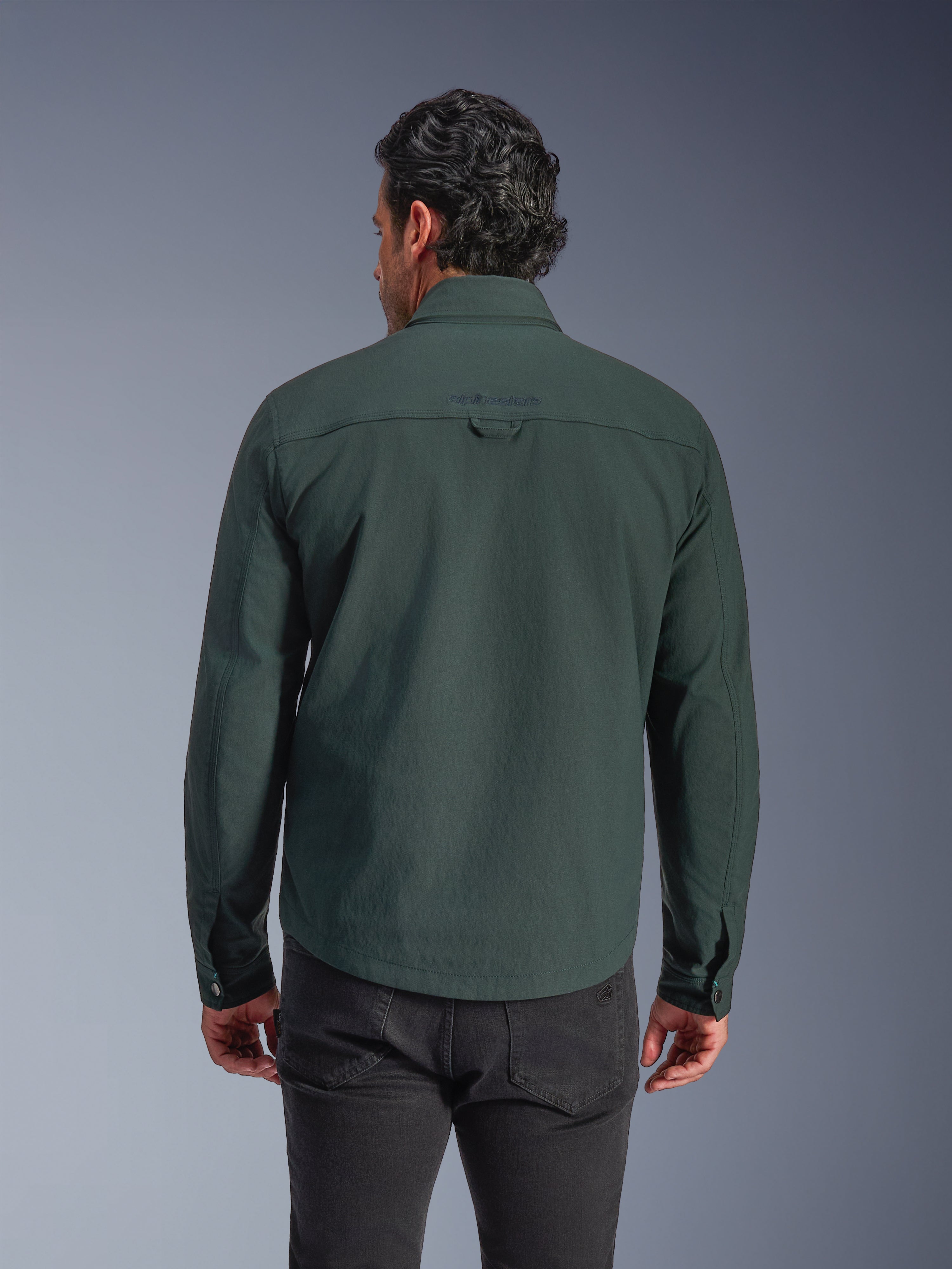 Alpinestars Bryon Water Resistant Overshirt, verde esmeralda oscuro, vista trasera en modelo, logotipo bordado en la parte superior de la espalda, lona encerada duradera, chaqueta de moto urbana combinada con pantalones oscuros