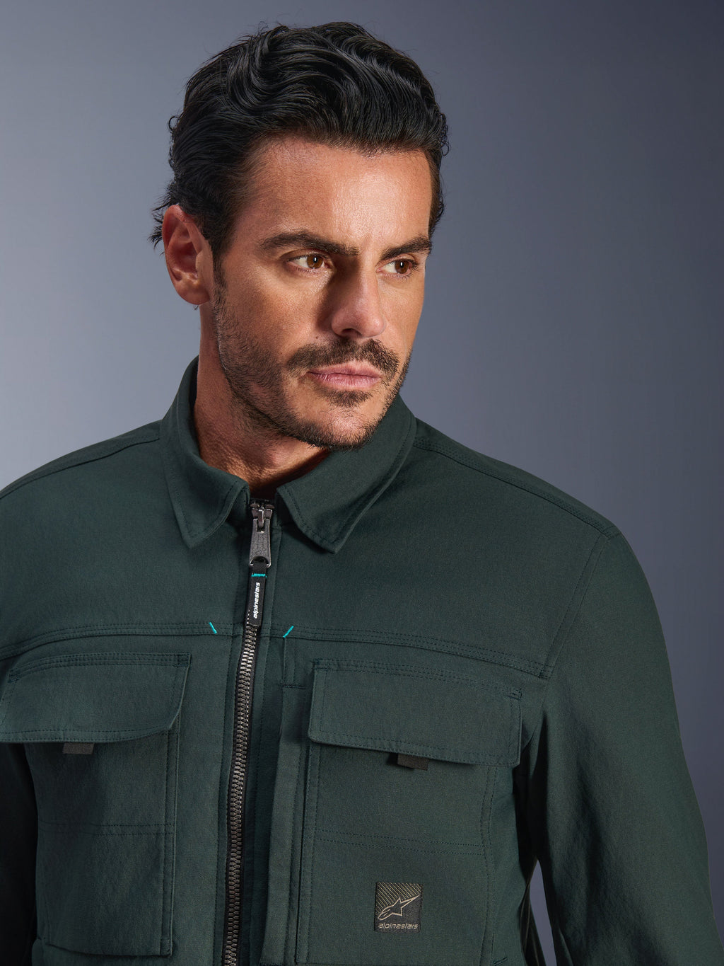 Alpinestars Bryon Water Resistant Overshirt en color Dark Emerald, retrato en primer plano de un hombre con esta chaqueta urbana de moto, con textura de lona encerada, cremallera frontal con sutiles detalles en verde azulado, bolsillos tipo cargo en el pecho y parche con el logotipo de la marca.