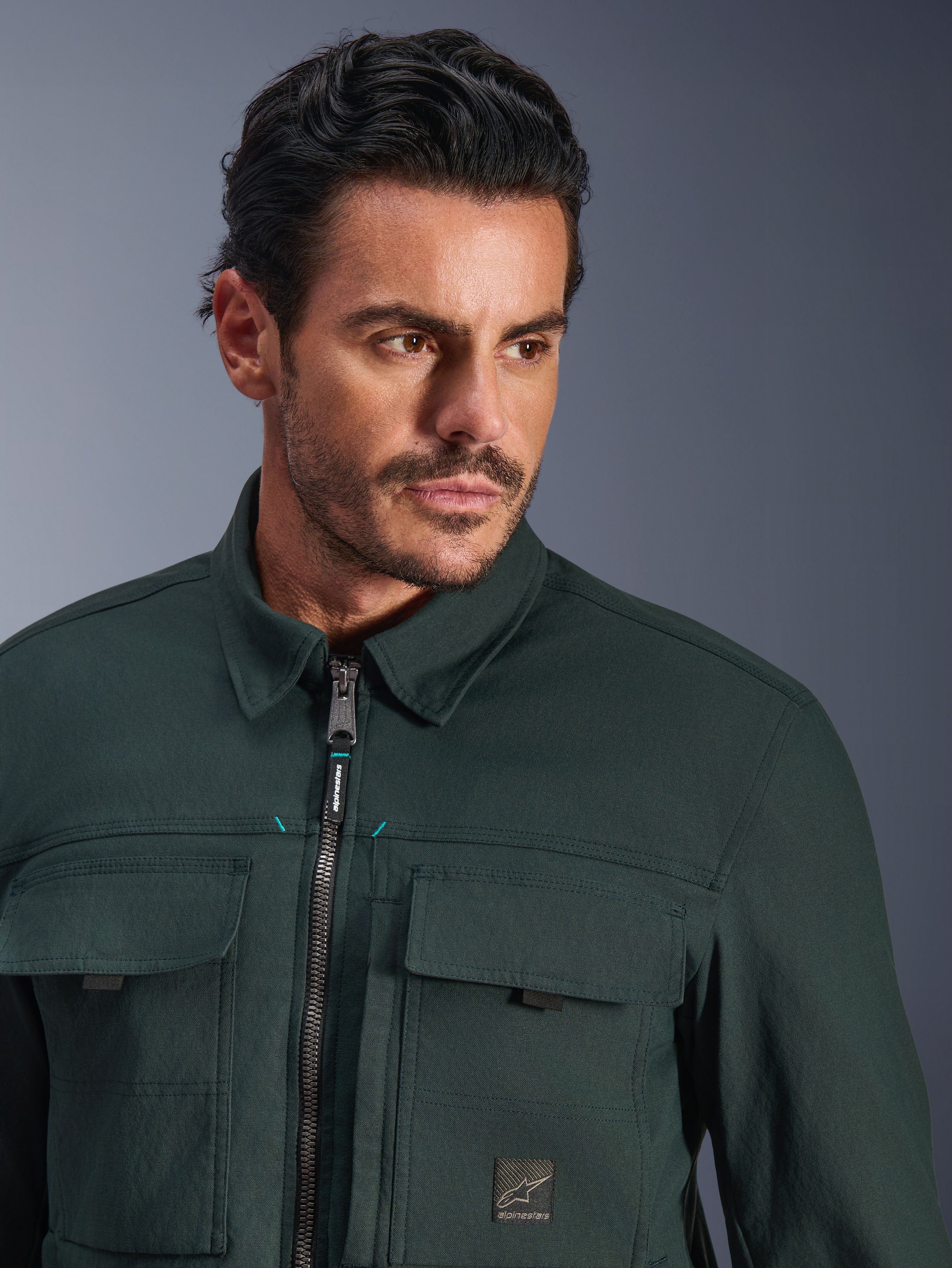 Alpinestars Bryon Water Resistant Overshirt en color Dark Emerald, retrato en primer plano de un hombre con esta chaqueta urbana de moto, con textura de lona encerada, cremallera frontal con sutiles detalles en verde azulado, bolsillos tipo cargo en el pecho y parche con el logotipo de la marca.