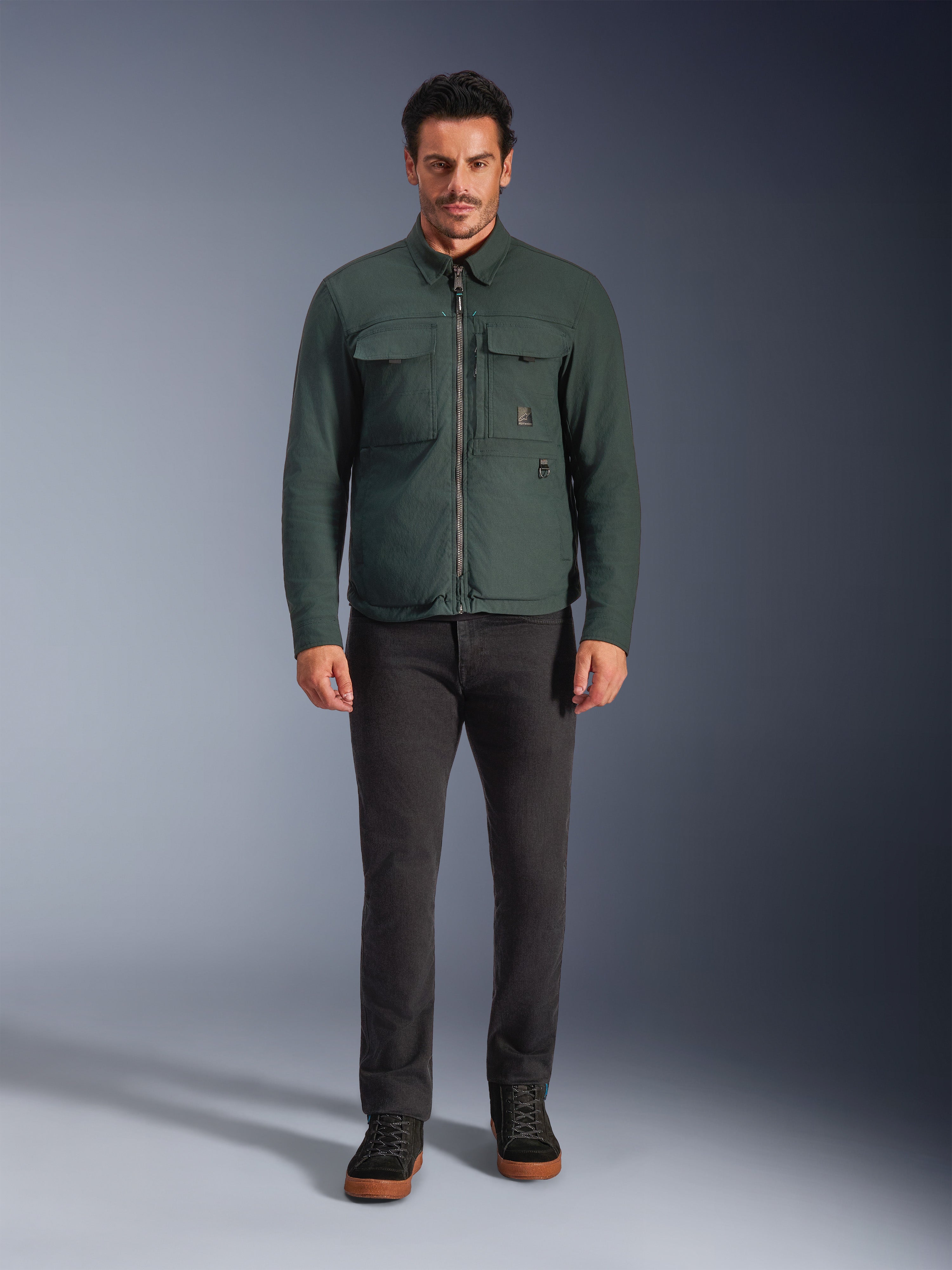 Sobrecamisa Alpinestars Bryon WR, chaqueta de moto resistente al agua, esmeralda oscuro, combinada con pantalones negros y botas negras, con confección en lona encerada, bolsillos cargo en el pecho y un diseño urbano moderno