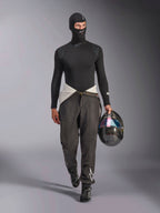 Alpinestars ZX Evo V3 Balaclava, negro, usado por un modelo con un conjunto completo de capas base para carreras, que incluye una parte superior de manga larga a juego y pantalones de carreras grises, sosteniendo un casco de fibra de carbono con visera iridiscente, equipamiento profesional para automovilismo